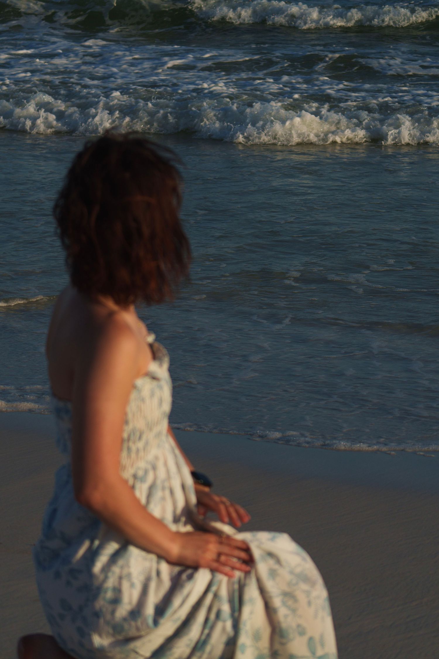 Young woman in blue floral dress sitting on sandy beach watching ocean waves roll in at sunset.