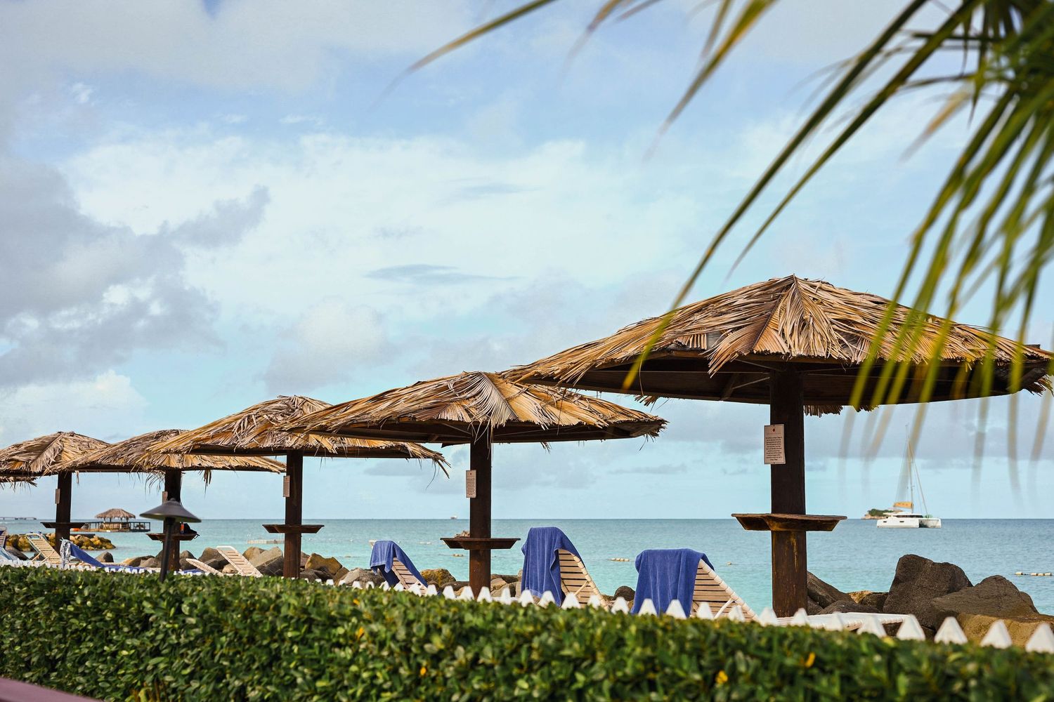 Beachfront area with thatched umbrellas, blue lounge chairs, and a sailboat on calm Caribbean waters.