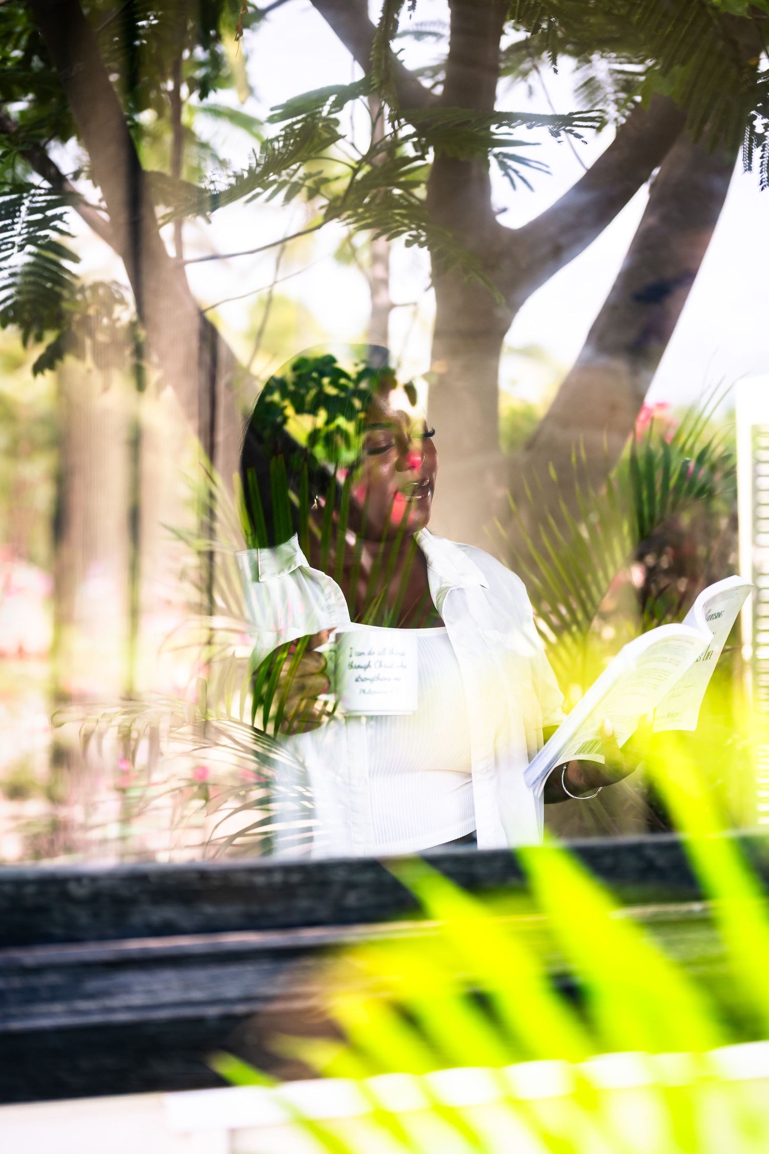 Woman in white shirt holding a mug, viewed through glass with tropical garden reflection.