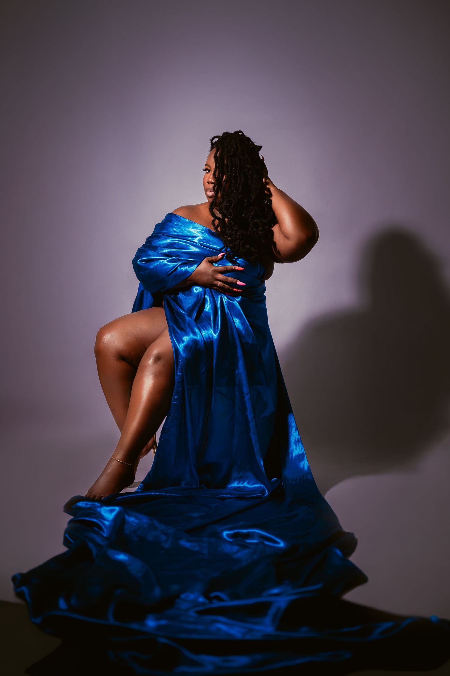 A plus-size Black woman poses gracefully in a flowing royal blue satin off-shoulder gown against a soft purple backdrop.