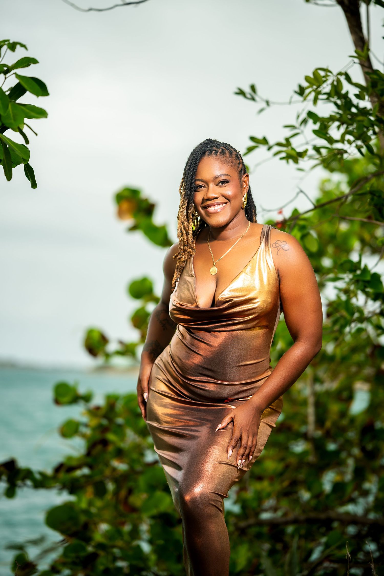 Smiling woman in gold satin dress posing among tropical trees with ocean in background.