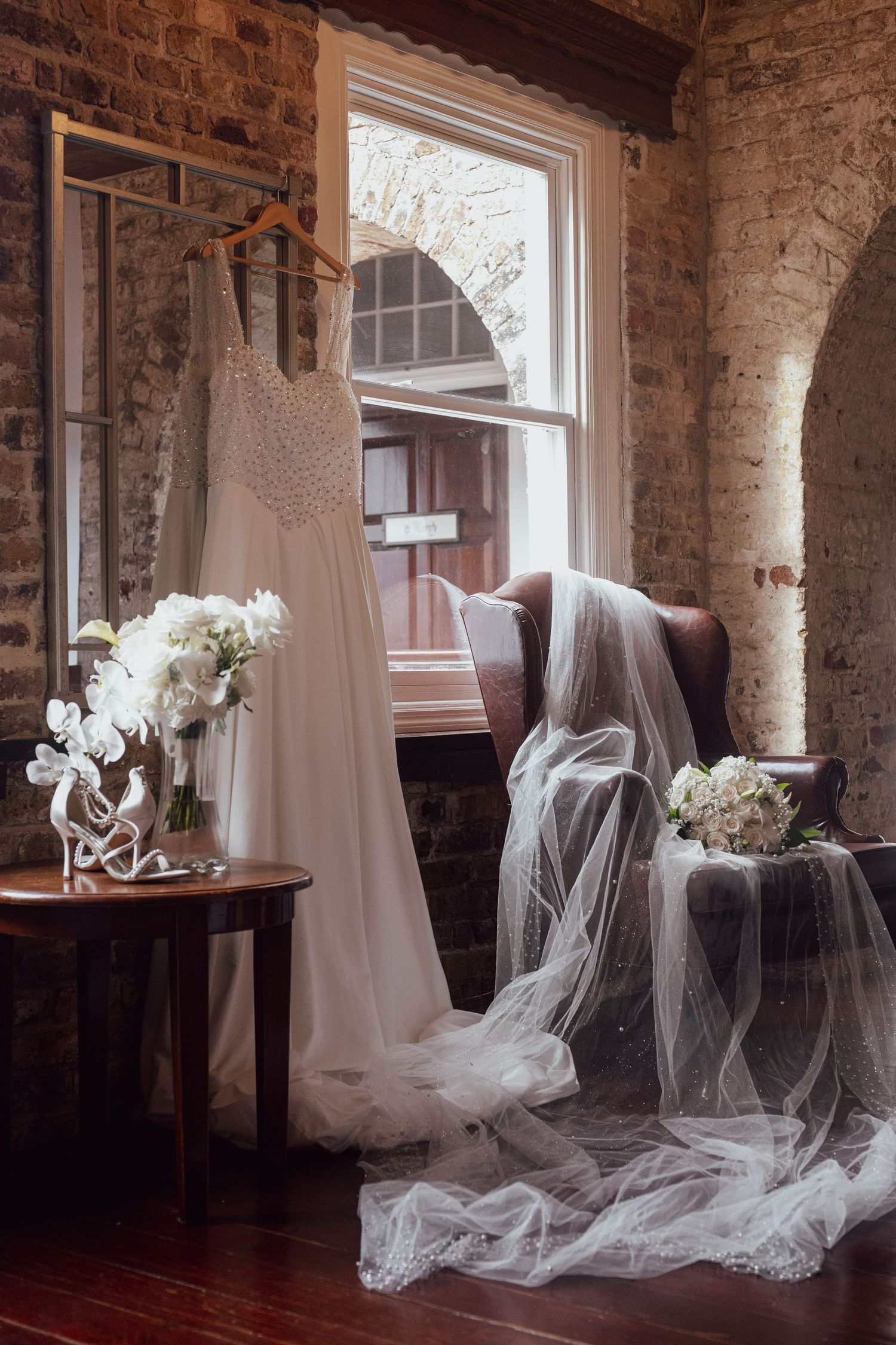 Elegant wedding dress hanging near window with bridal veil draped over armchair and white floral bouquets in rustic brick room.