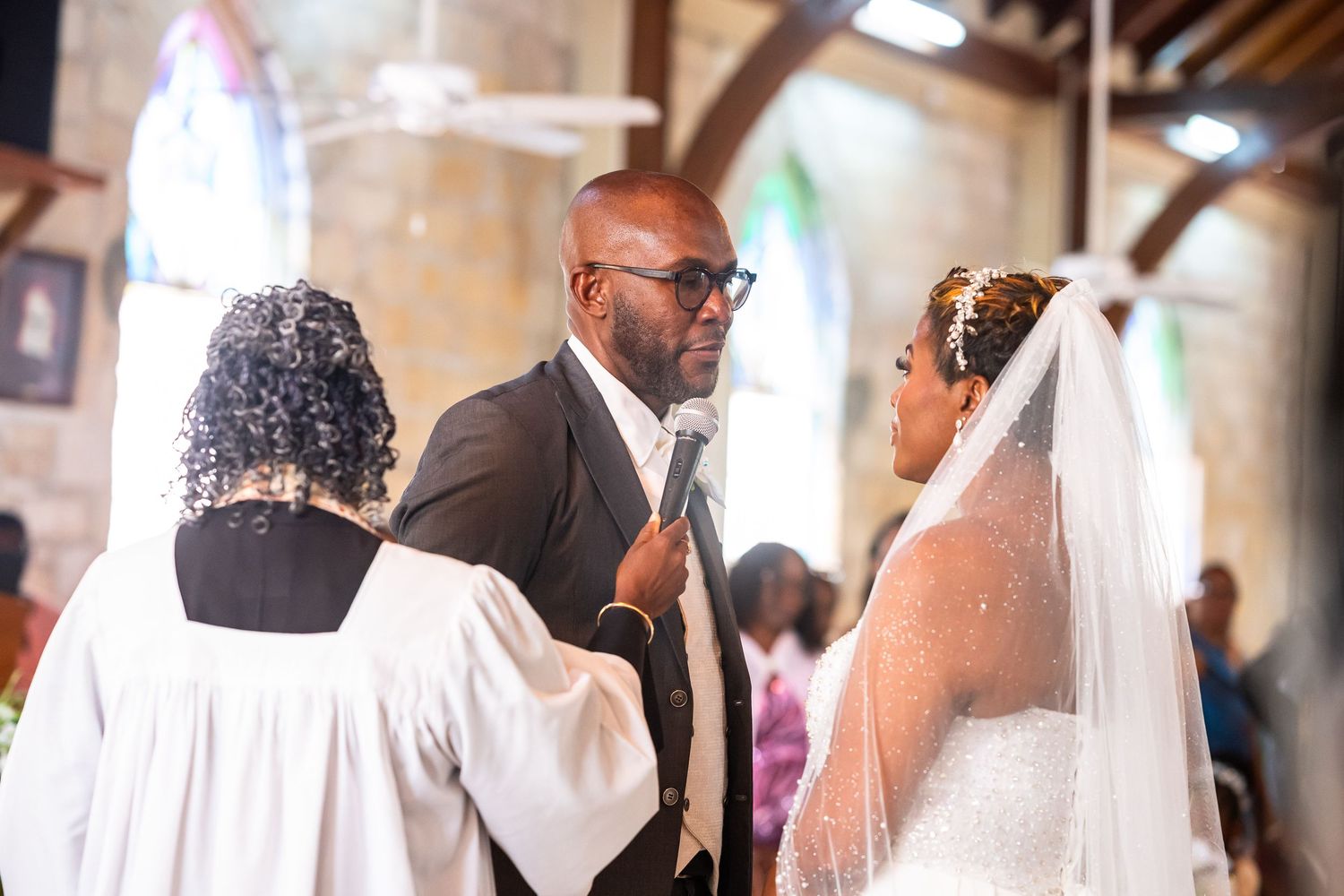 A groom in a gray suit exchanges vows with his bride in a white gown and veil during a church wedding ceremony.
