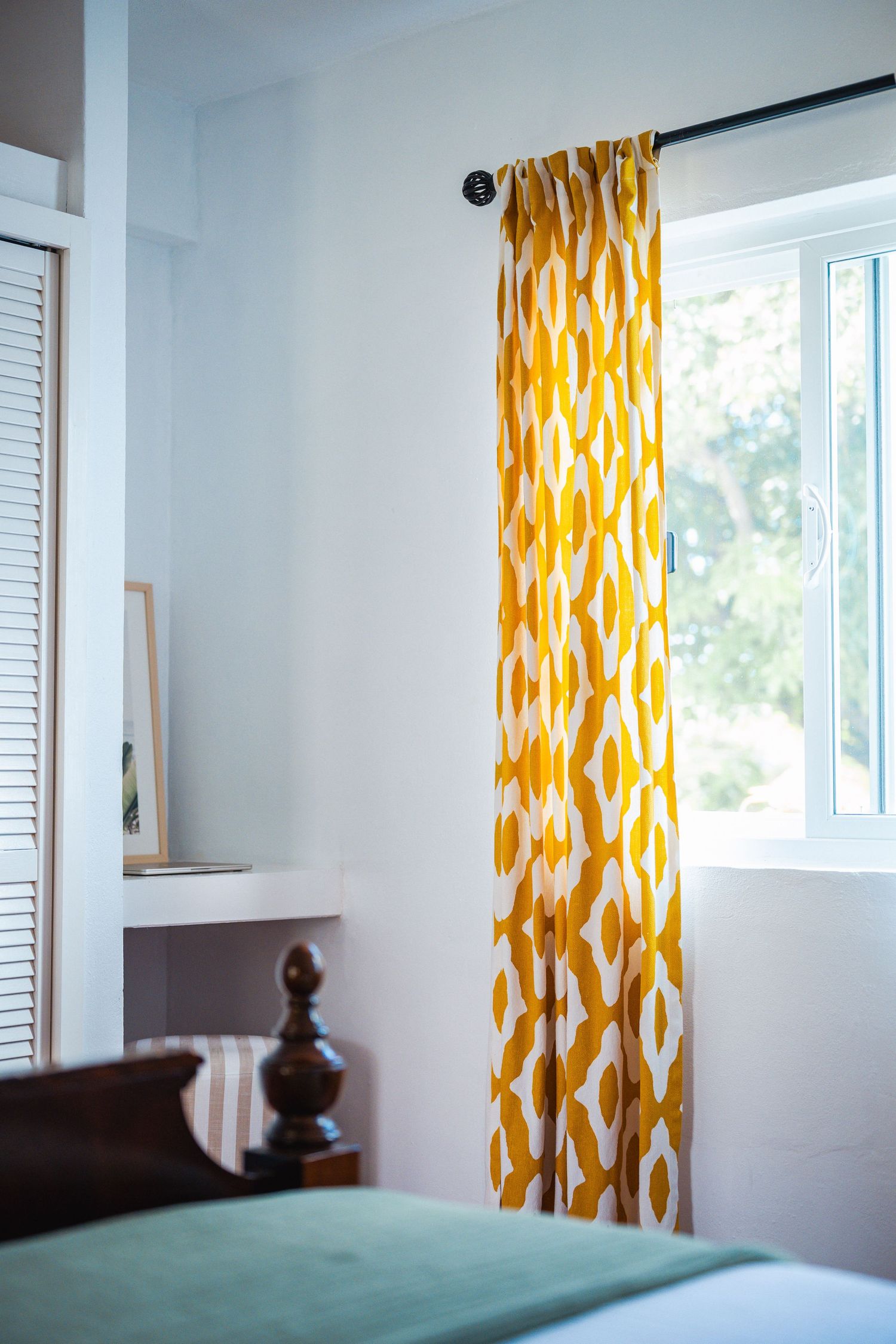Bright bedroom with bold yellow geometric patterned curtains beside a white window with tropical garden views.