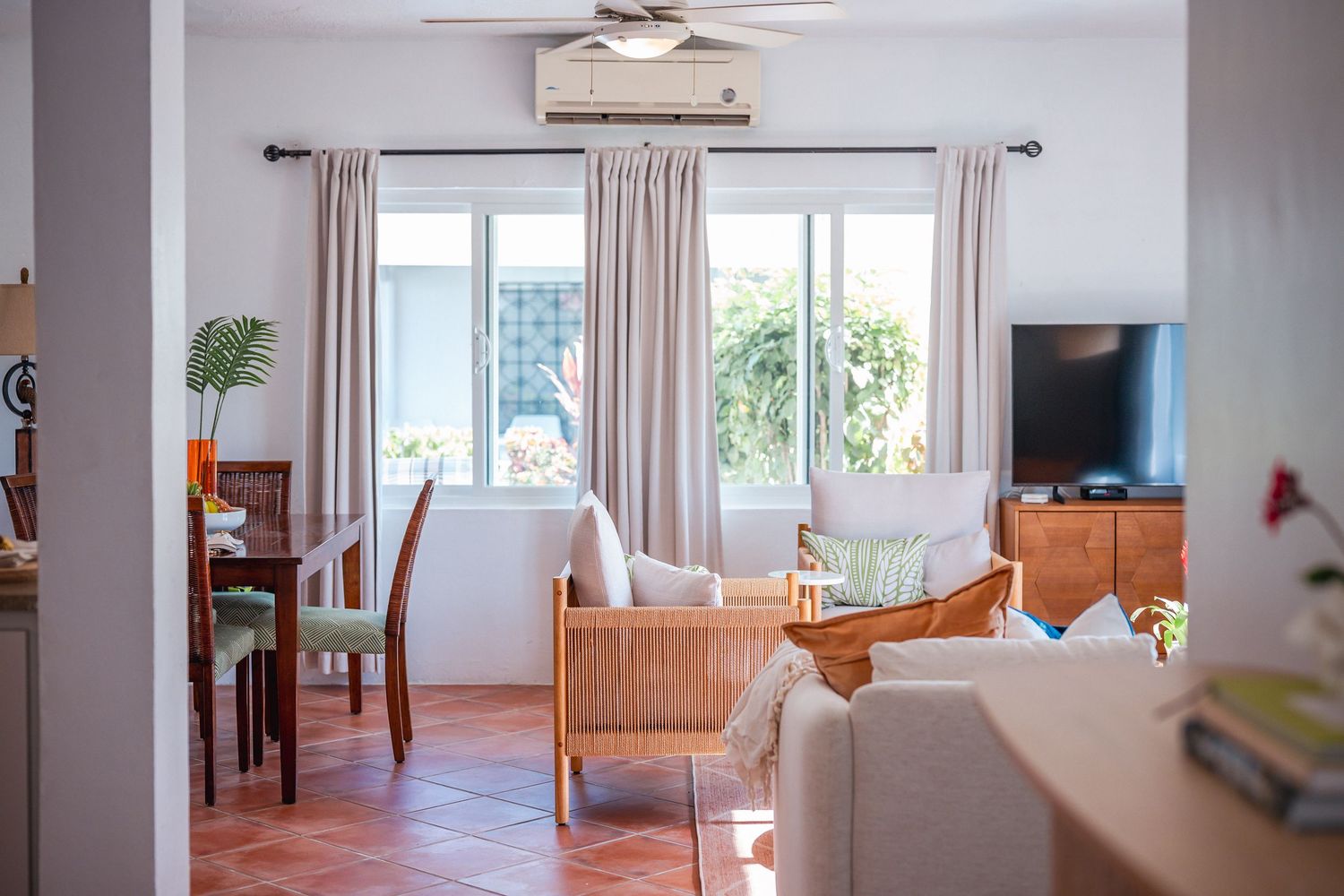 Sunlit living room with wicker furniture, beige curtains, ceiling fan, and TV in a tropical villa.