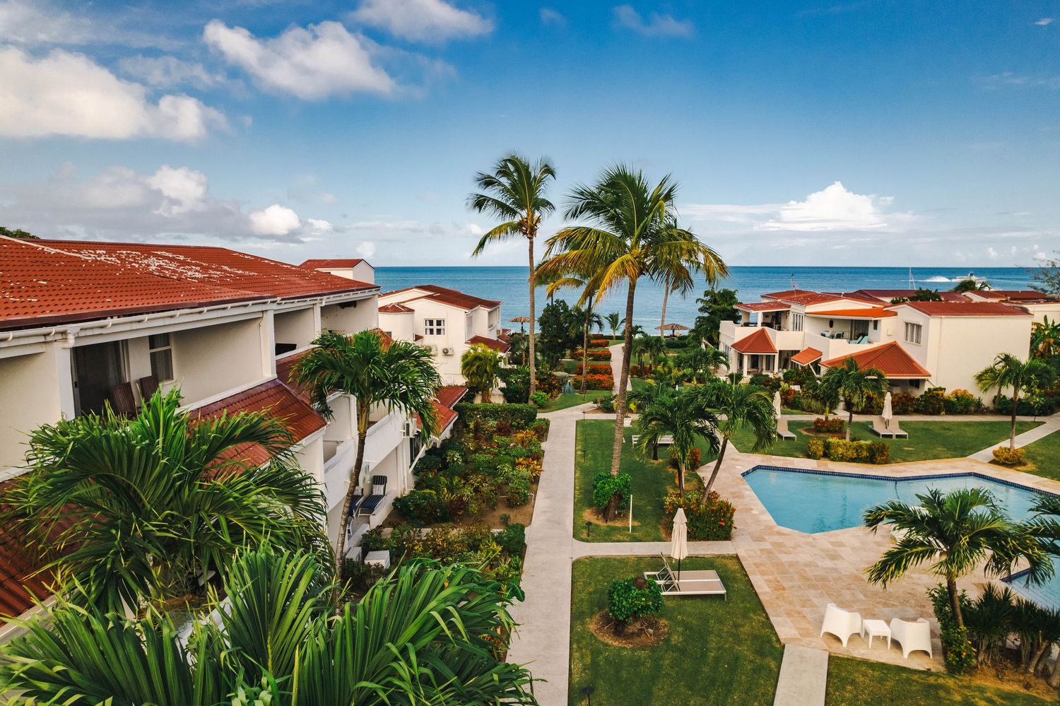 Aerial view of a tropical resort featuring white villas, manicured gardens, swimming pool, and ocean views.