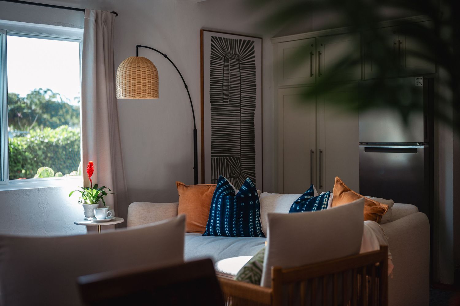 Modern tropical living room with rattan lamp, colorful pillows, and lush window view.