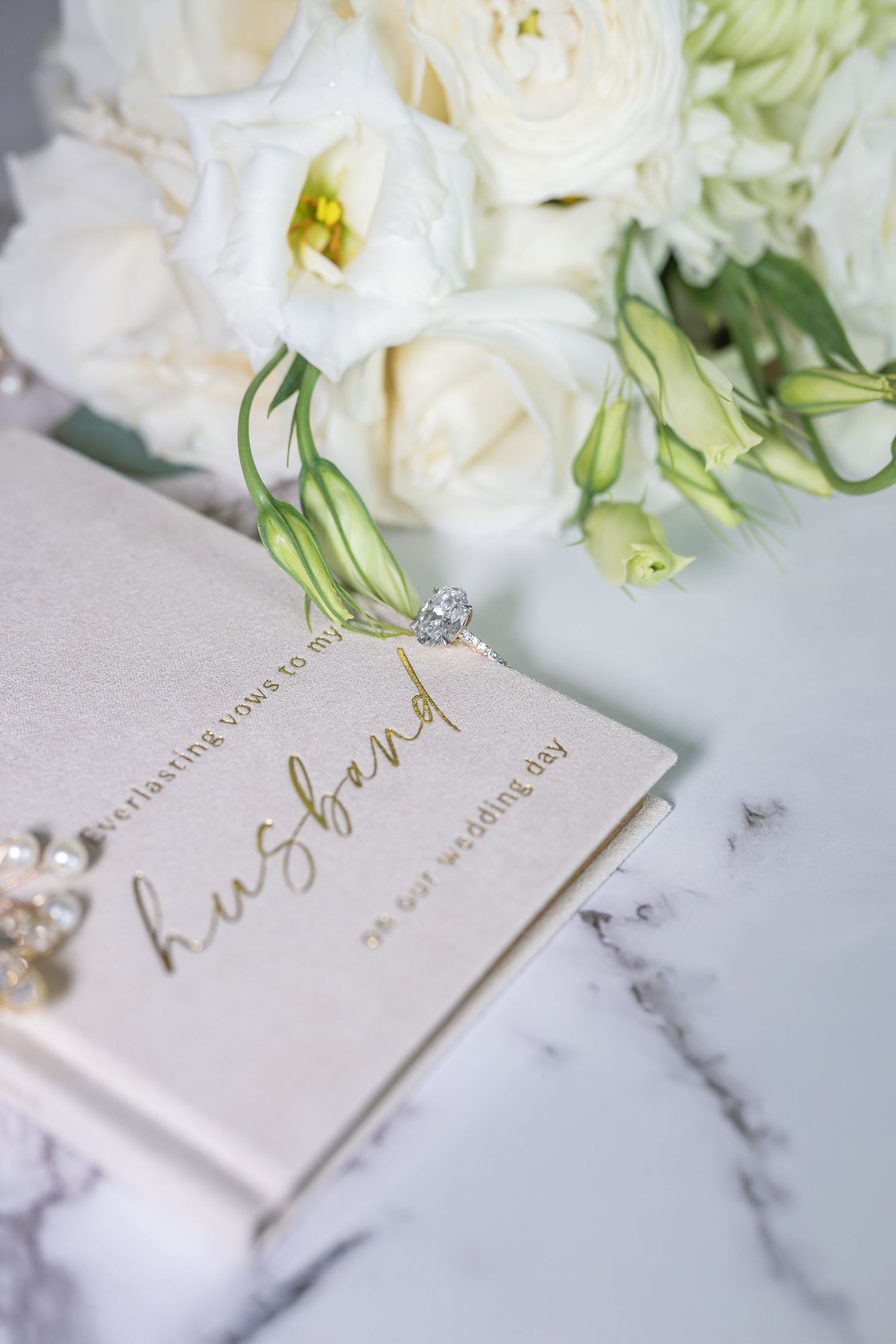 Elegant wedding flat lay with a husband vow book, diamond ring, pearl bracelet, and white floral bouquet on marble.