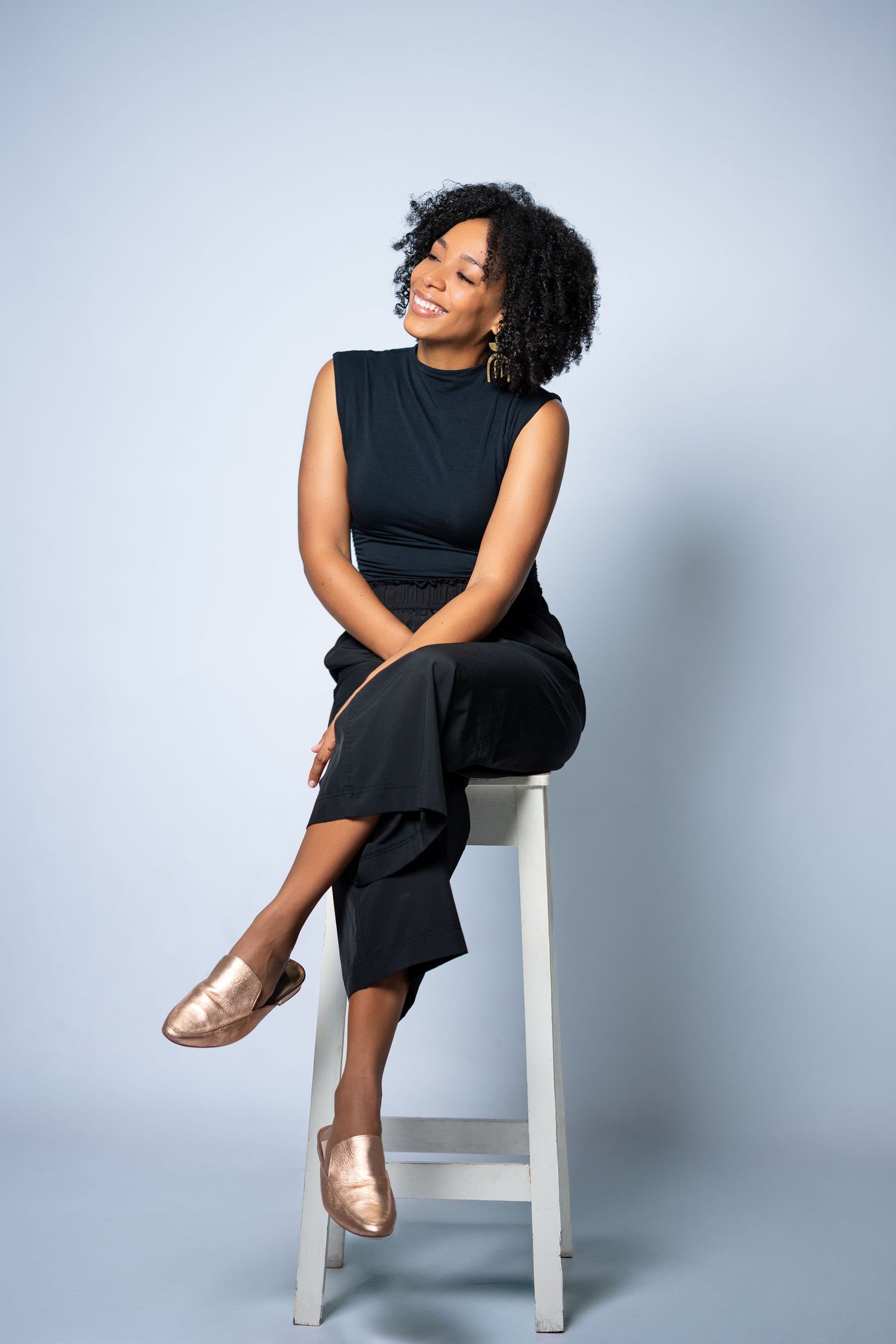 Smiling Black woman in black outfit and gold flats sitting on white stool against light blue background.