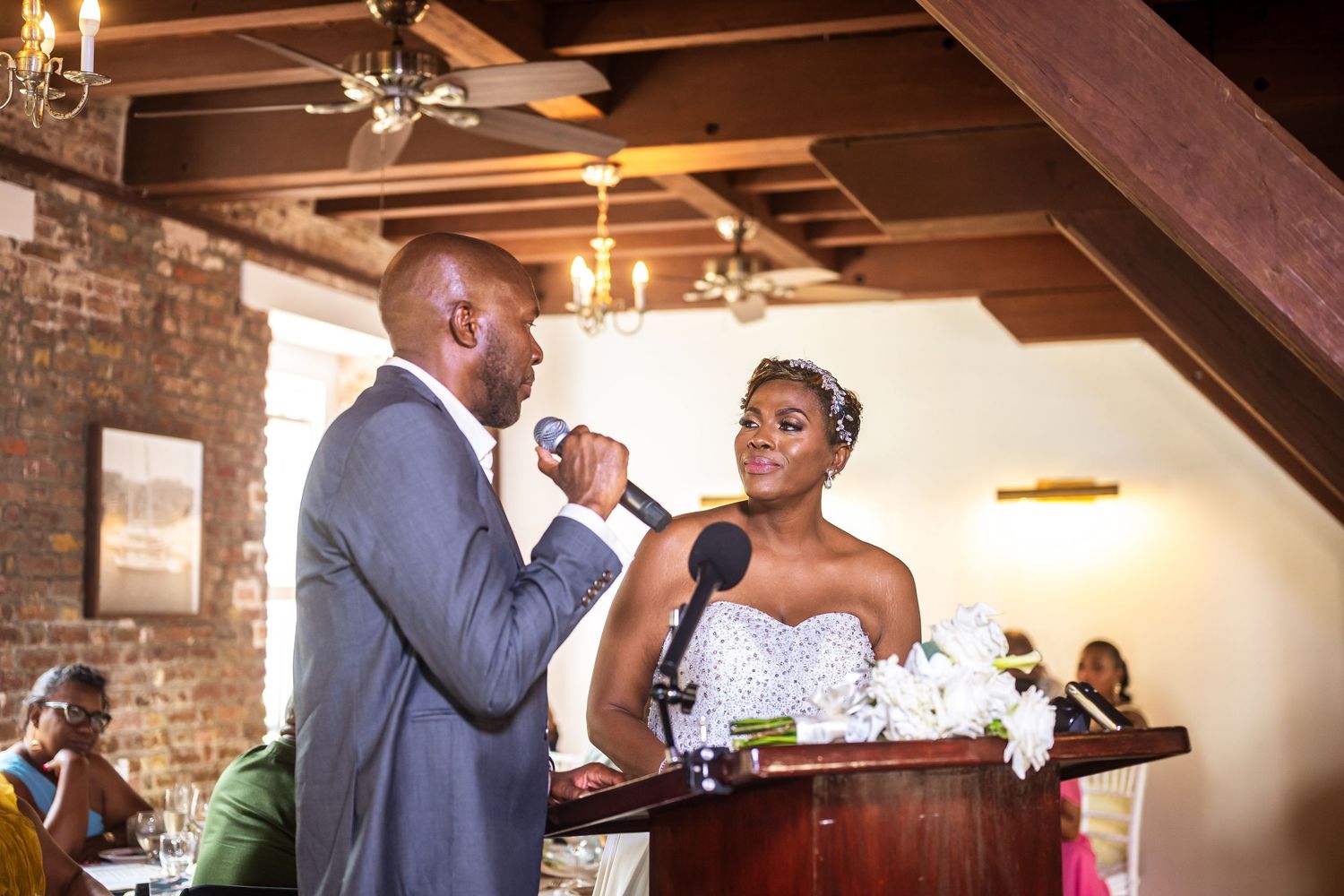 A groom in a gray suit speaks into a microphone beside his bride in a beaded strapless gown at their wedding reception.