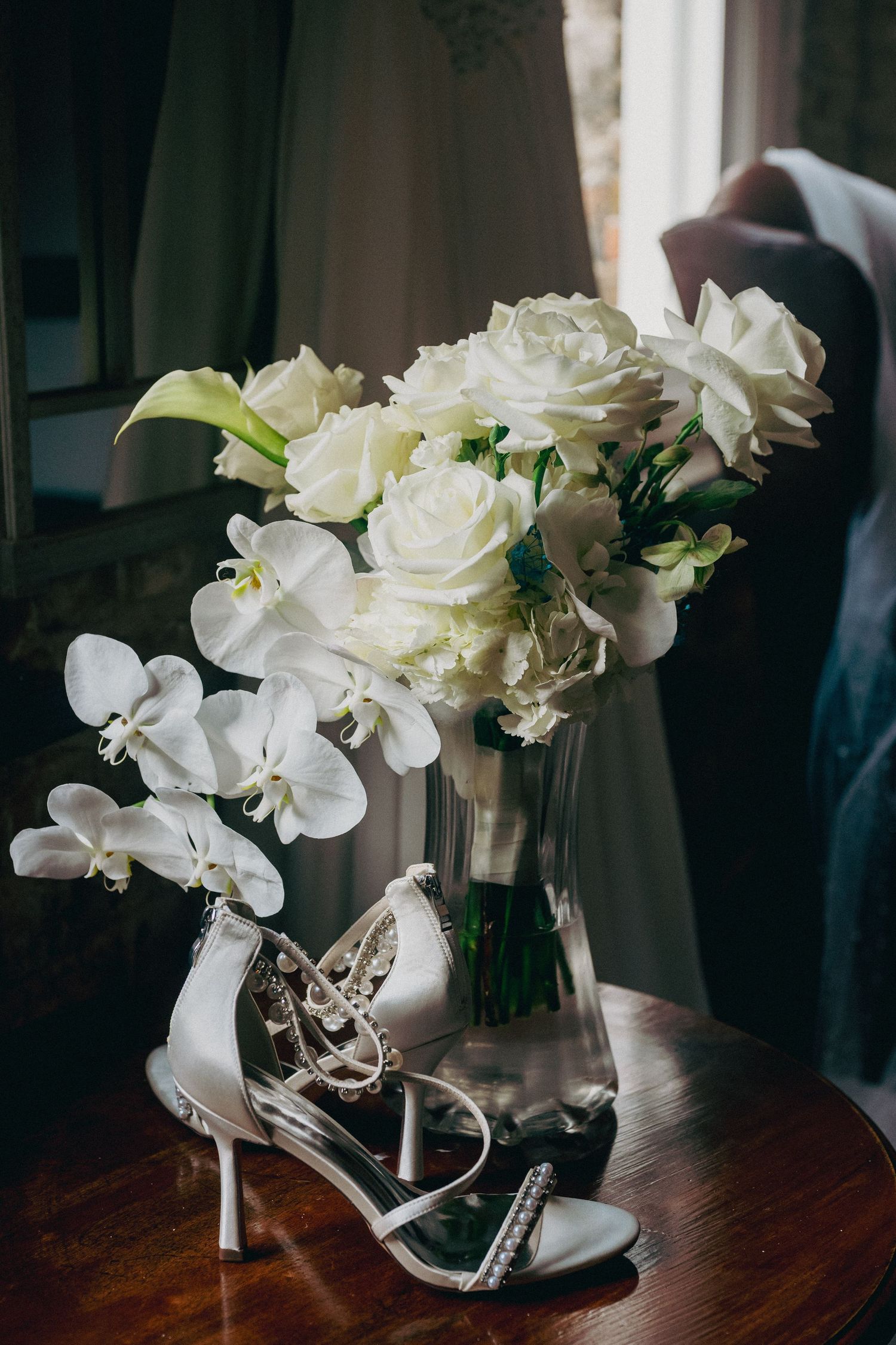 Elegant bridal bouquet of white roses and orchids beside crystal embellished wedding heels on a wooden table.