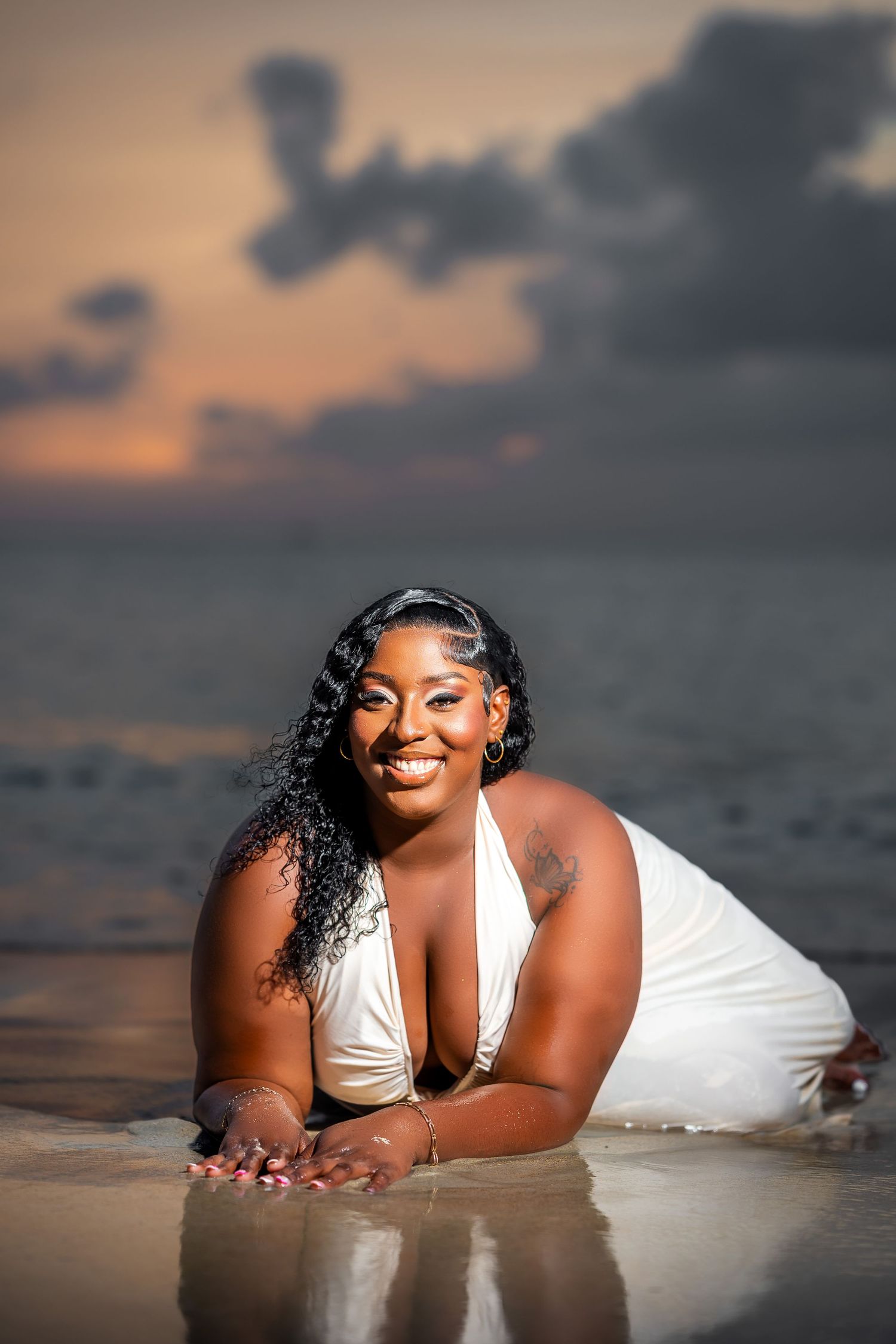 A smiling woman in a white halter dress poses on a wet beach at sunset, lying gracefully with the ocean behind her.