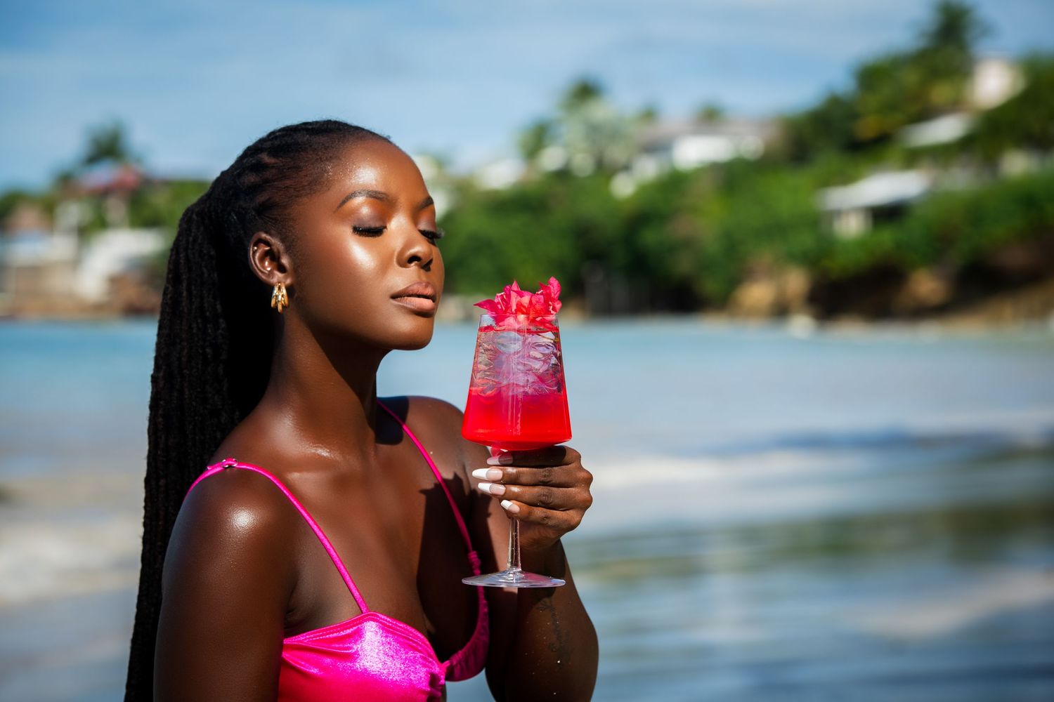 Young Black woman in pink bikini holding red cocktail on tropical beach with clear blue water and lush greenery.