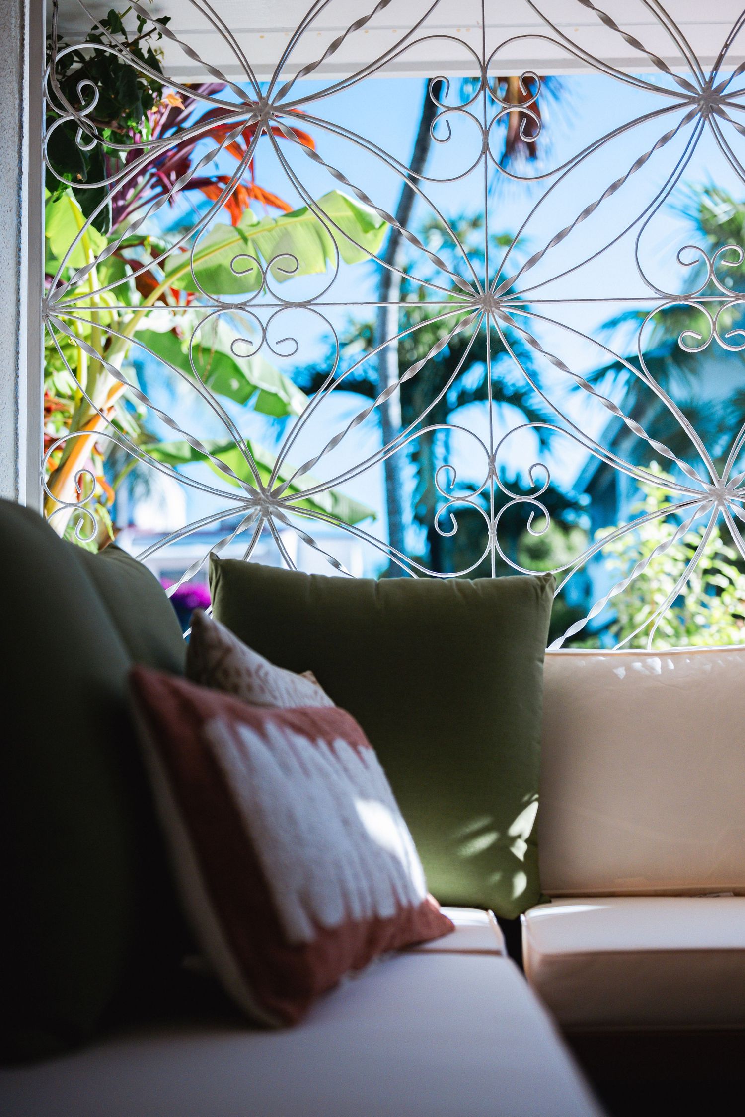 Close-up of beige sofa with green and patterned pillows against ornate wrought iron window with tropical garden backdrop.