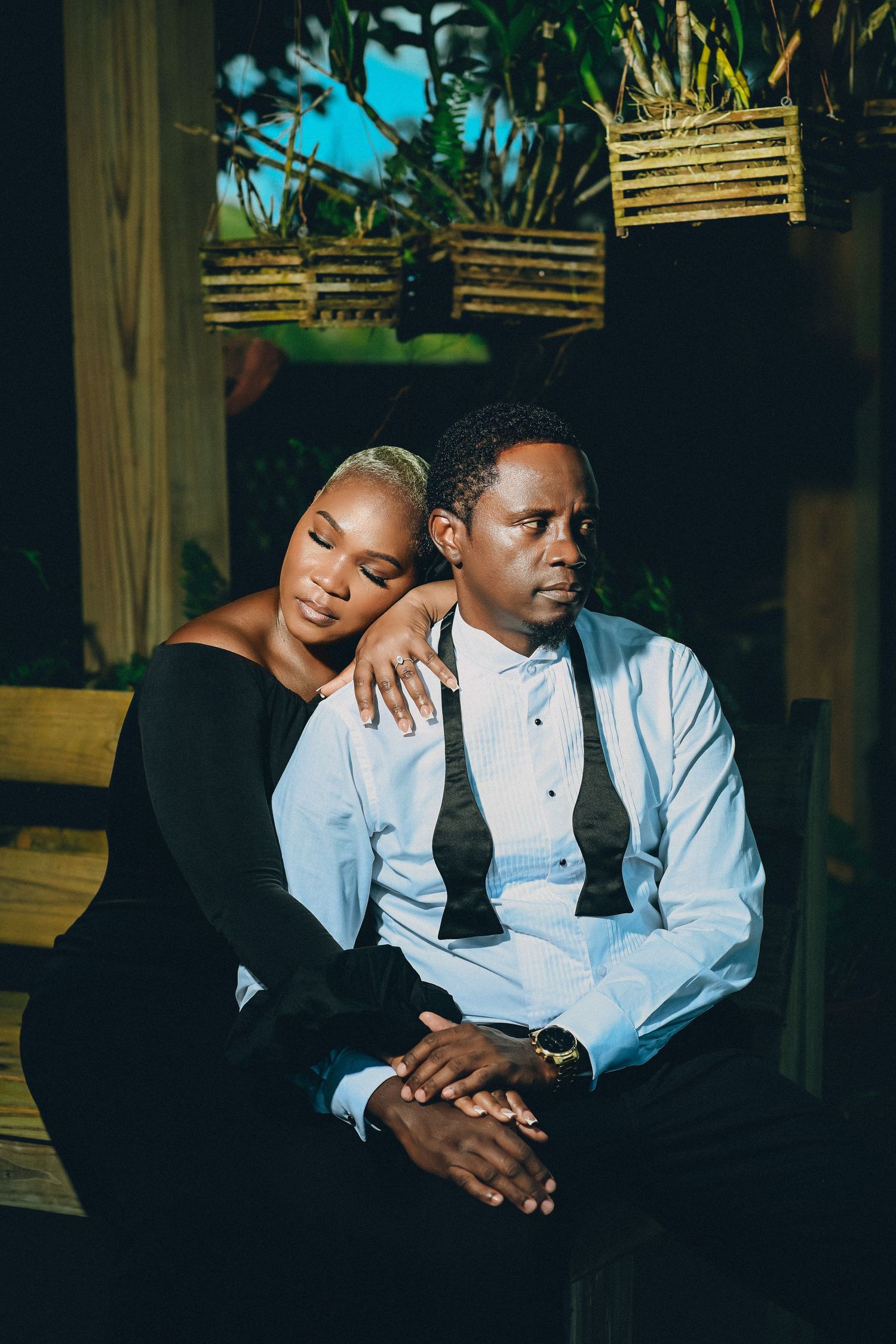 Elegant Black couple posing together in a dark romantic setting with tropical plants.