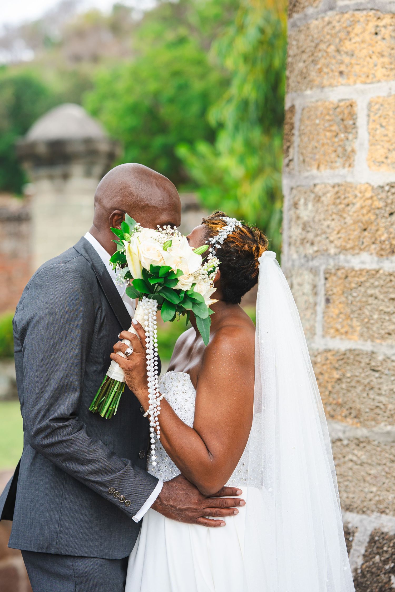 A newlywed couple shares a kiss behind a white bouquet, bride in veil and gown, groom in gray suit, outdoors.