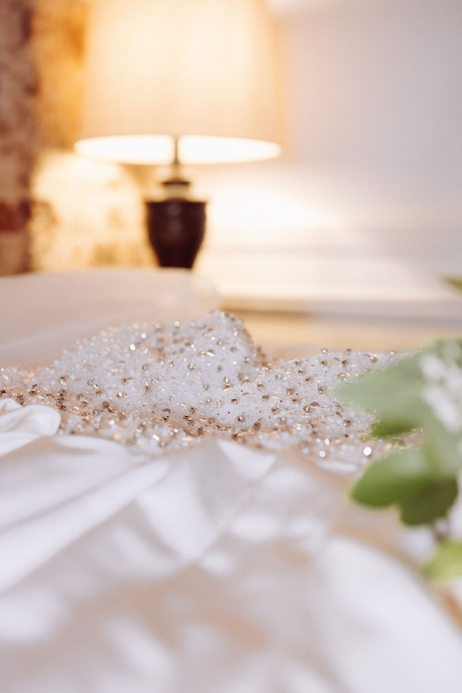 White wedding dress with pearl beading displayed on bed with glowing lamp in background.