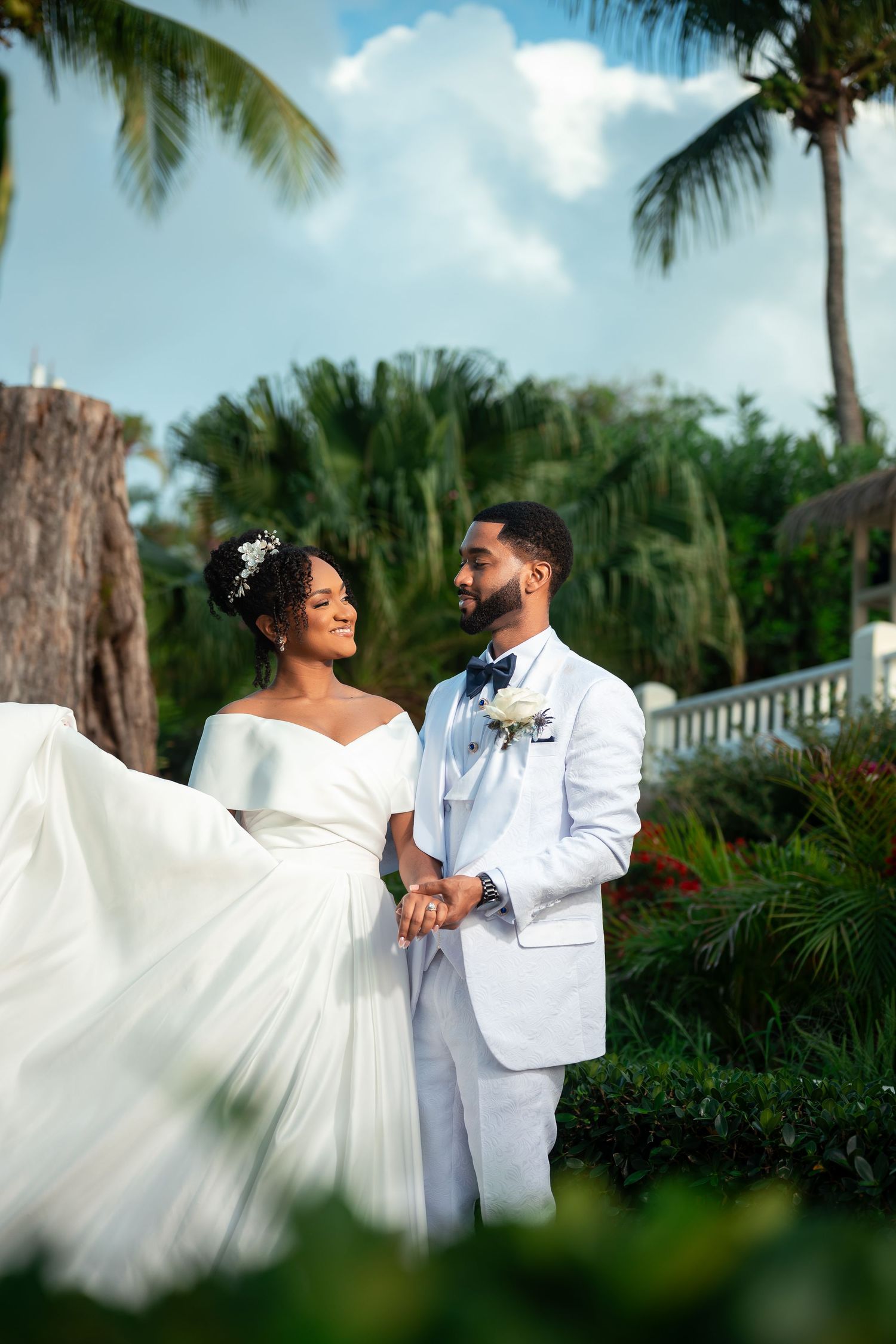 Bride in white off-shoulder gown and groom in white suit hold hands outdoors among tropical palm trees.