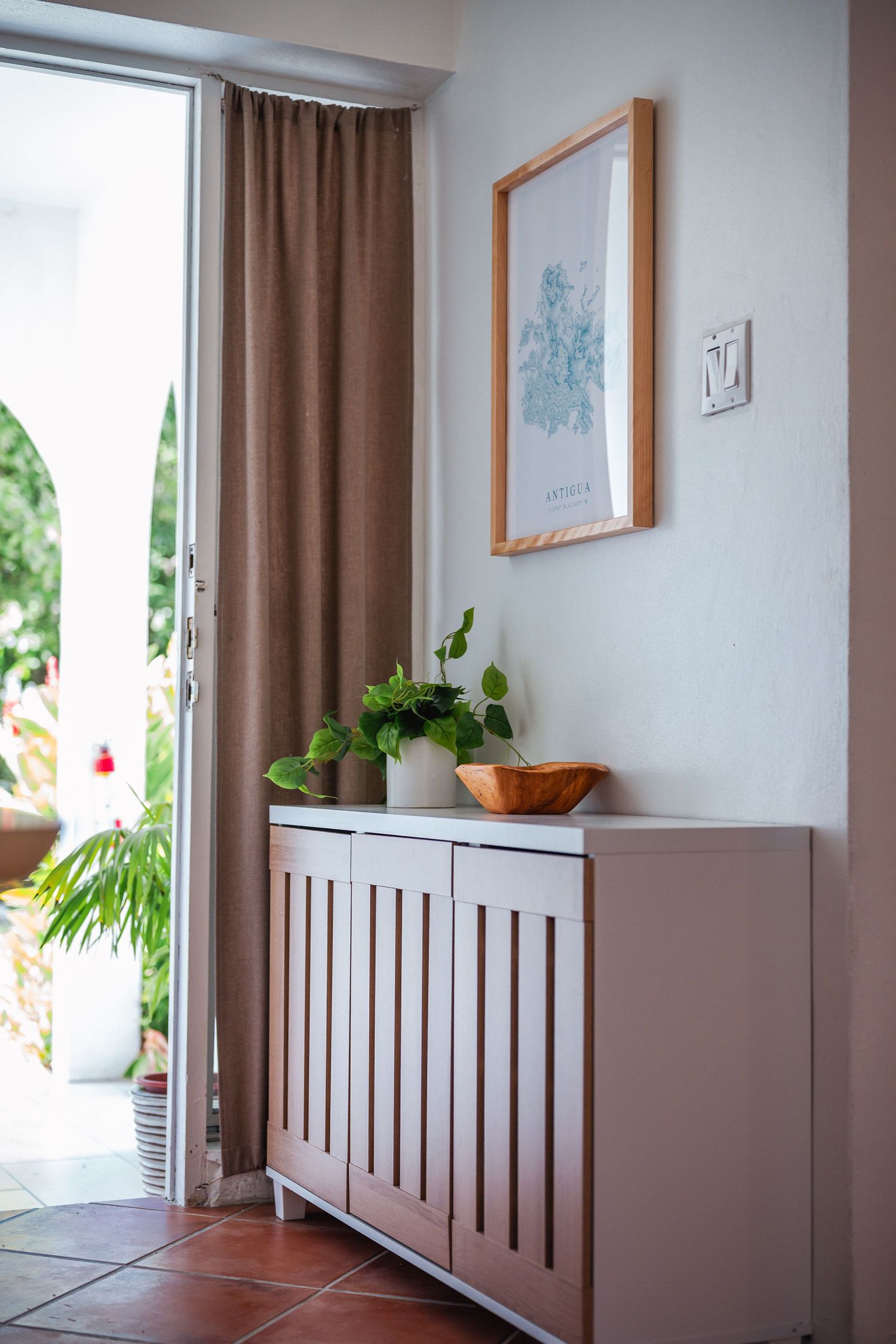 Entryway with pink cabinet, green potted plants, framed botanical art, and open door to tropical garden.