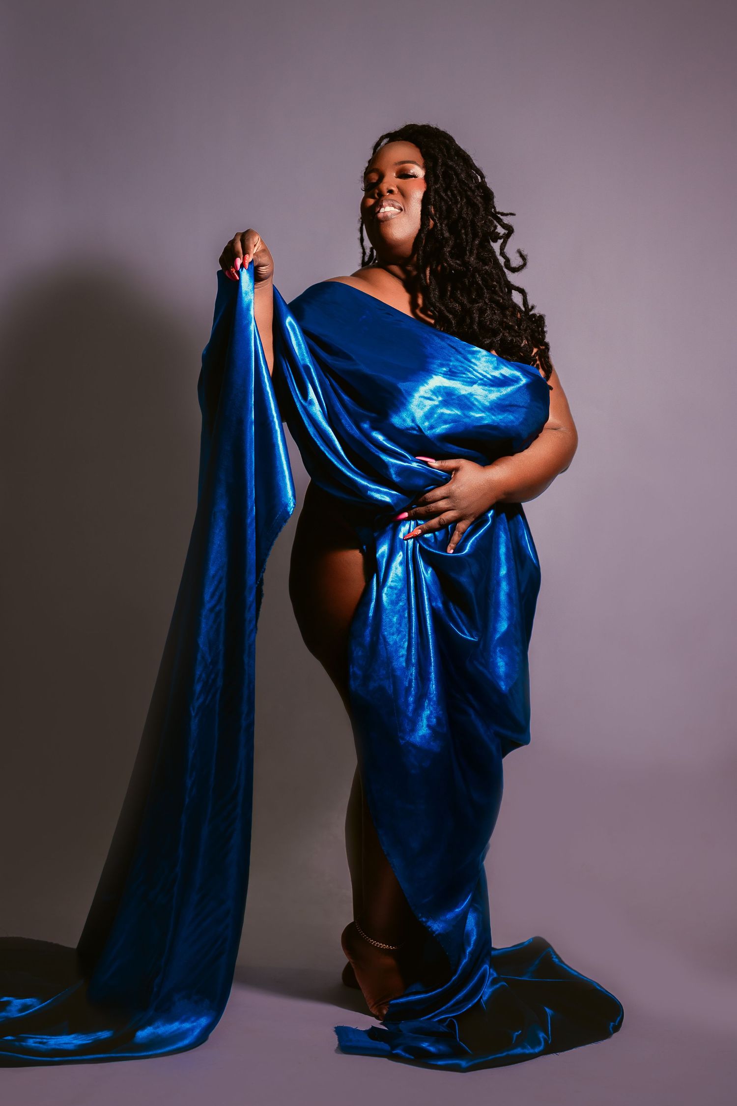 A plus-size woman poses elegantly draped in shiny royal blue satin fabric against a gray studio background.