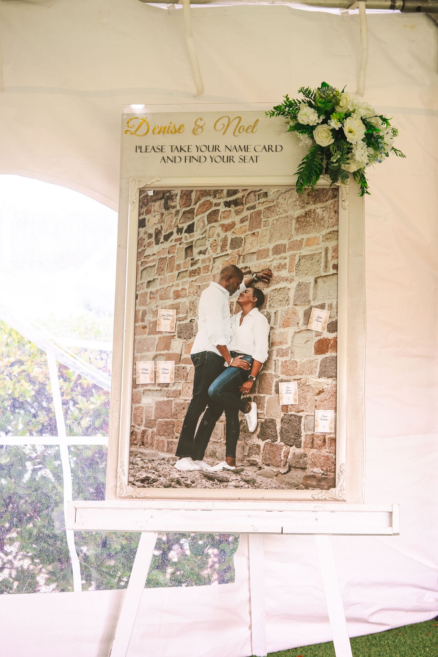 Wedding seating chart display featuring a couple's photo, floral arrangement, and name cards on an easel at a reception.