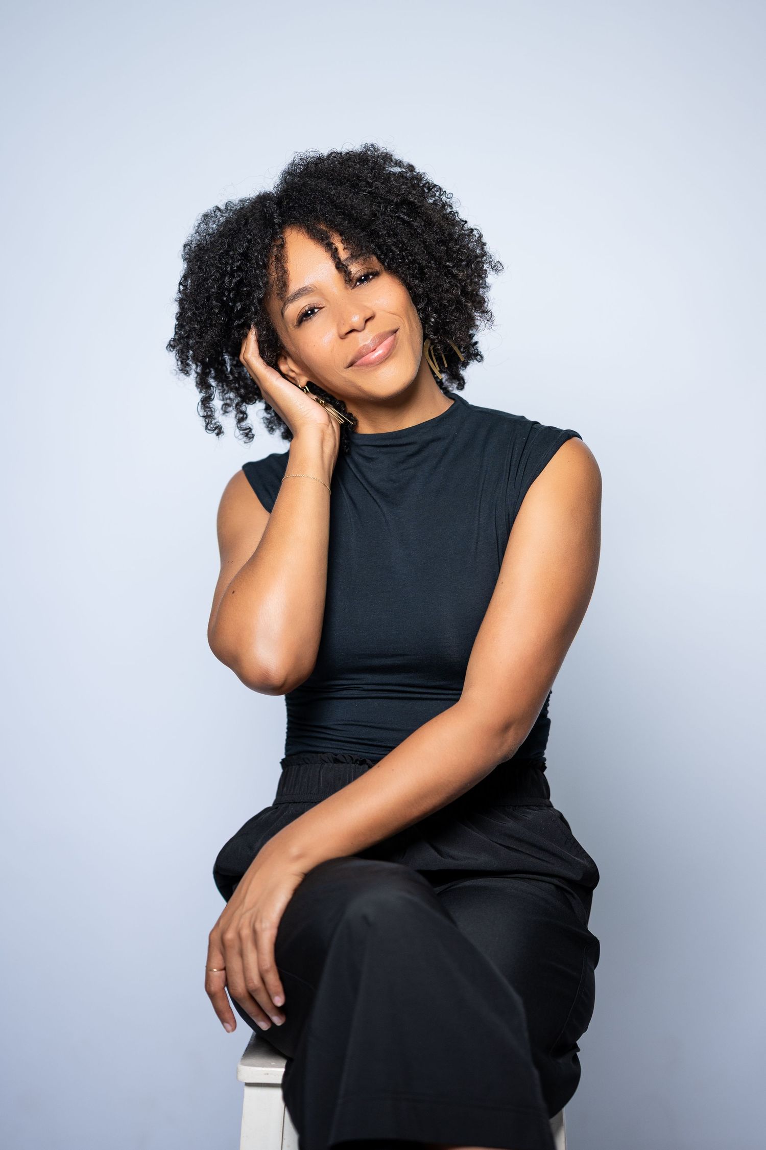 A young Black woman with curly hair sits gracefully in a black outfit against a light blue background.