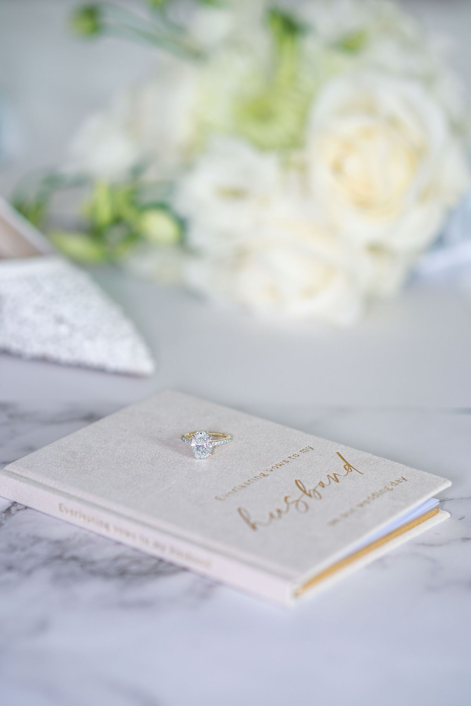Diamond engagement ring resting on a gold-lettered husband card, with white roses bouquet and silver heels in background.