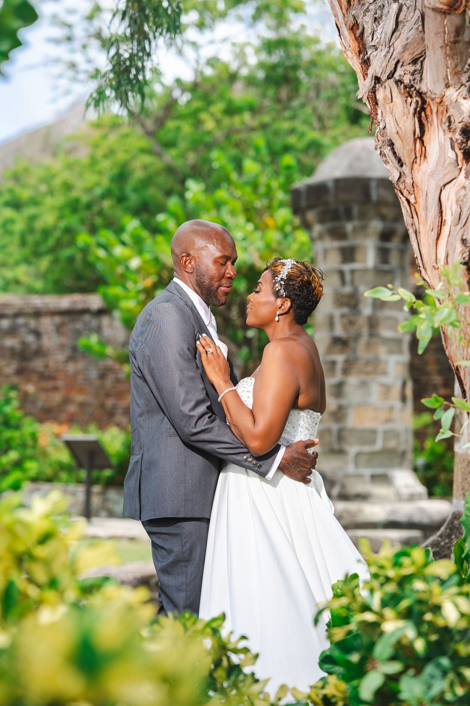 A bride and groom share a romantic moment outdoors, surrounded by lush greenery and stone ruins.
