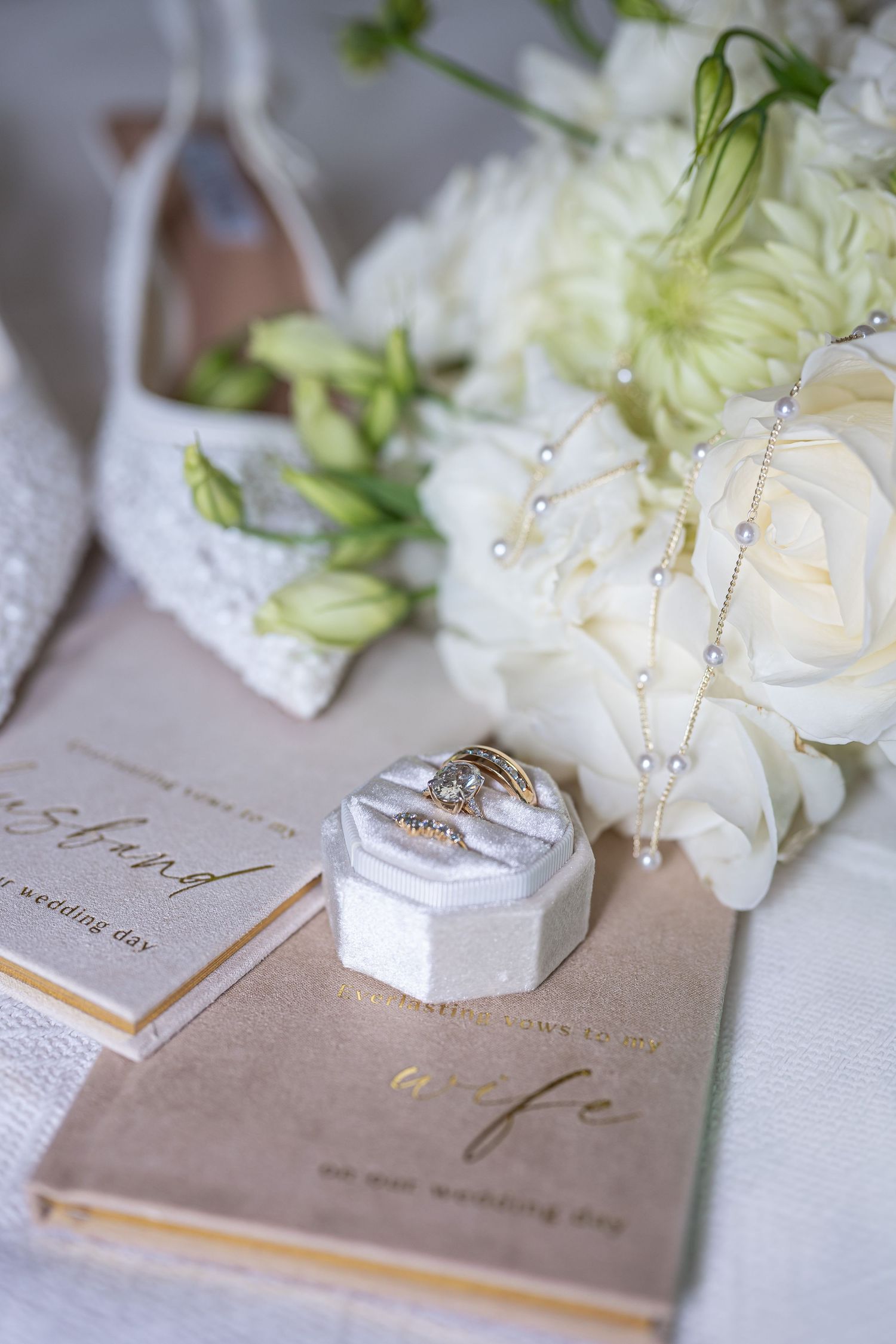 Elegant wedding flat lay with gold rings in velvet box, white floral bouquet, pearl necklace, lace heels, and vow books.