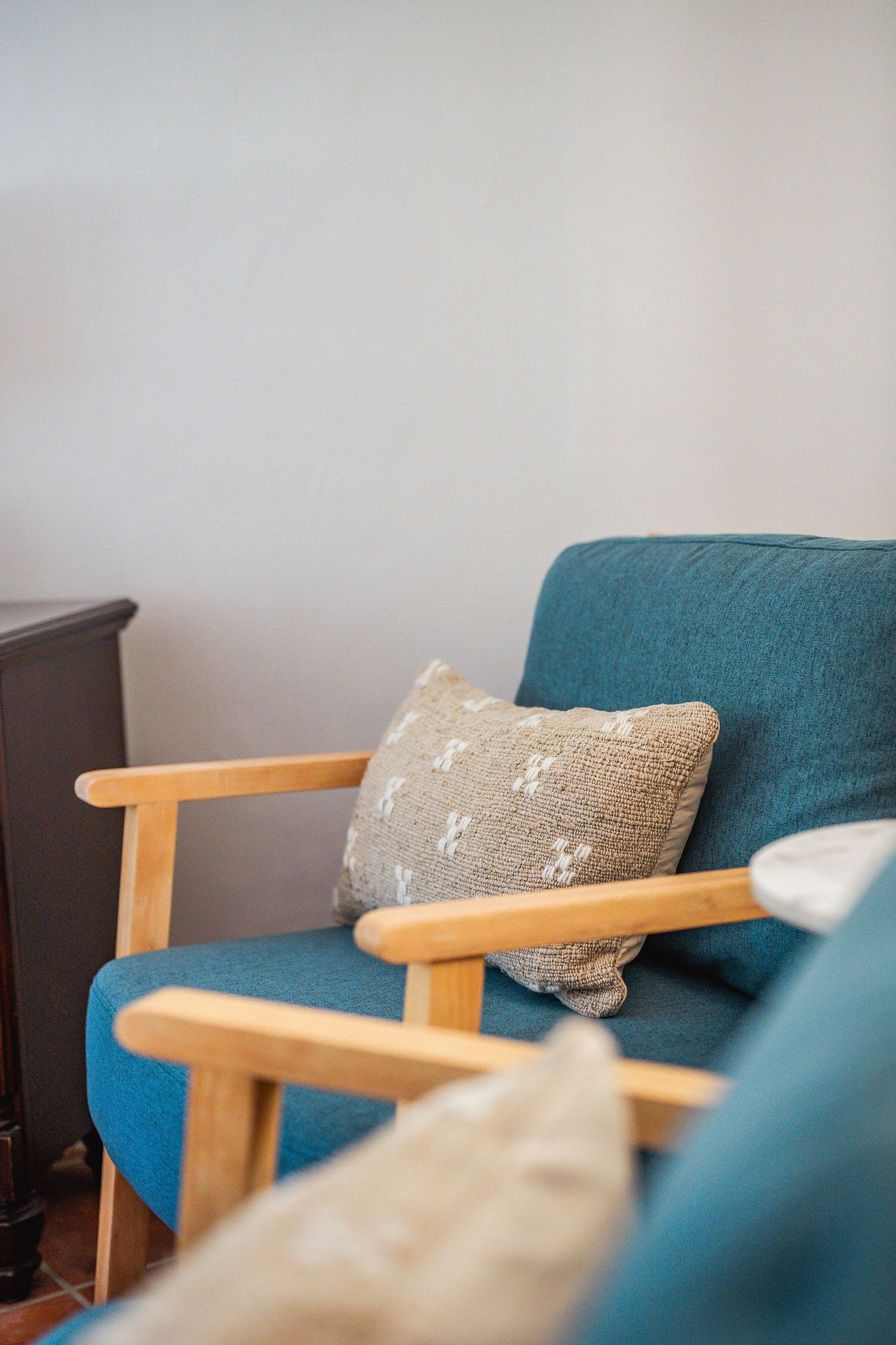 Teal mid-century modern armchair with wooden arms and neutral textured throw pillow against a clean white wall.