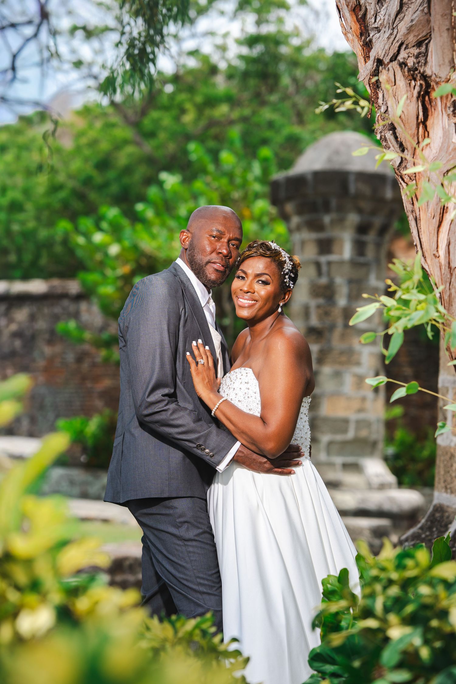 A happy couple in wedding attire poses outdoors near stone walls and lush greenery on their special day.