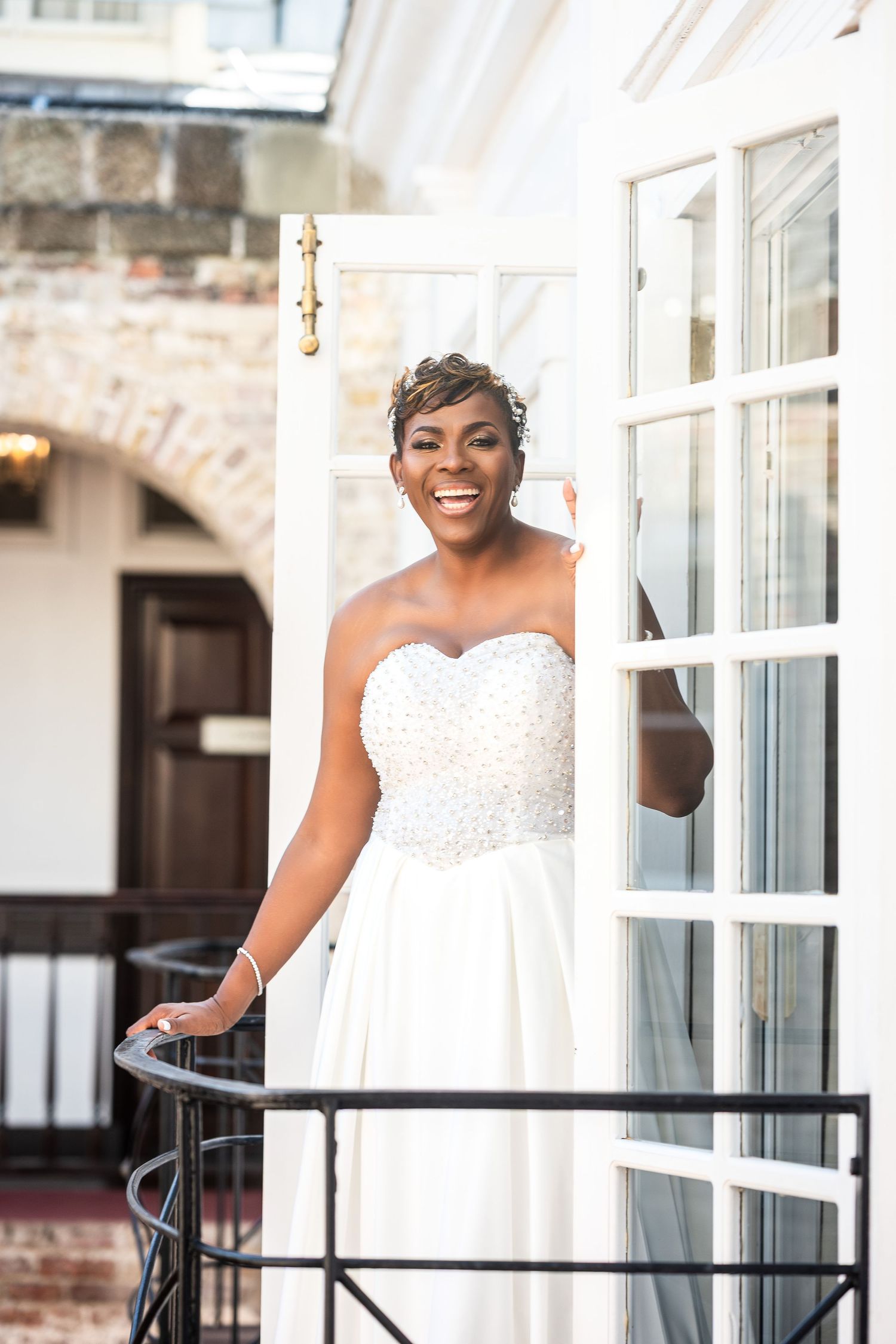 Smiling bride in strapless pearl wedding gown standing on balcony doorway of elegant venue.