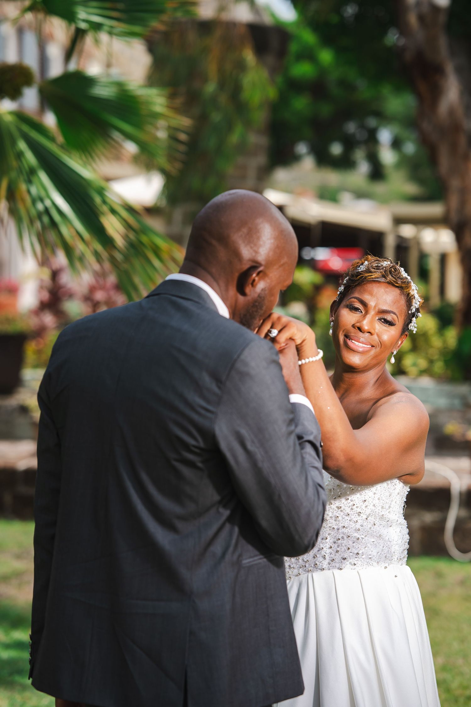 A joyful bride in a white gown smiles as groom kisses her hand during outdoor wedding ceremony.