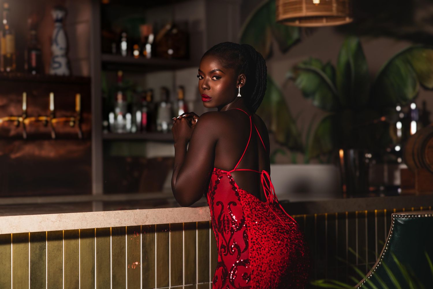 A woman in a red sequin dress poses elegantly at a bar, looking over her shoulder with dangling earrings.