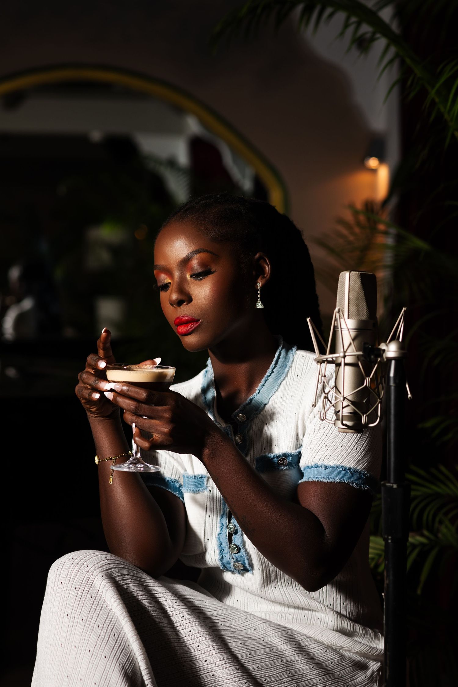 Young Black woman in white dress holds cocktail glass near studio microphone in moody, warmly lit recording studio setting.