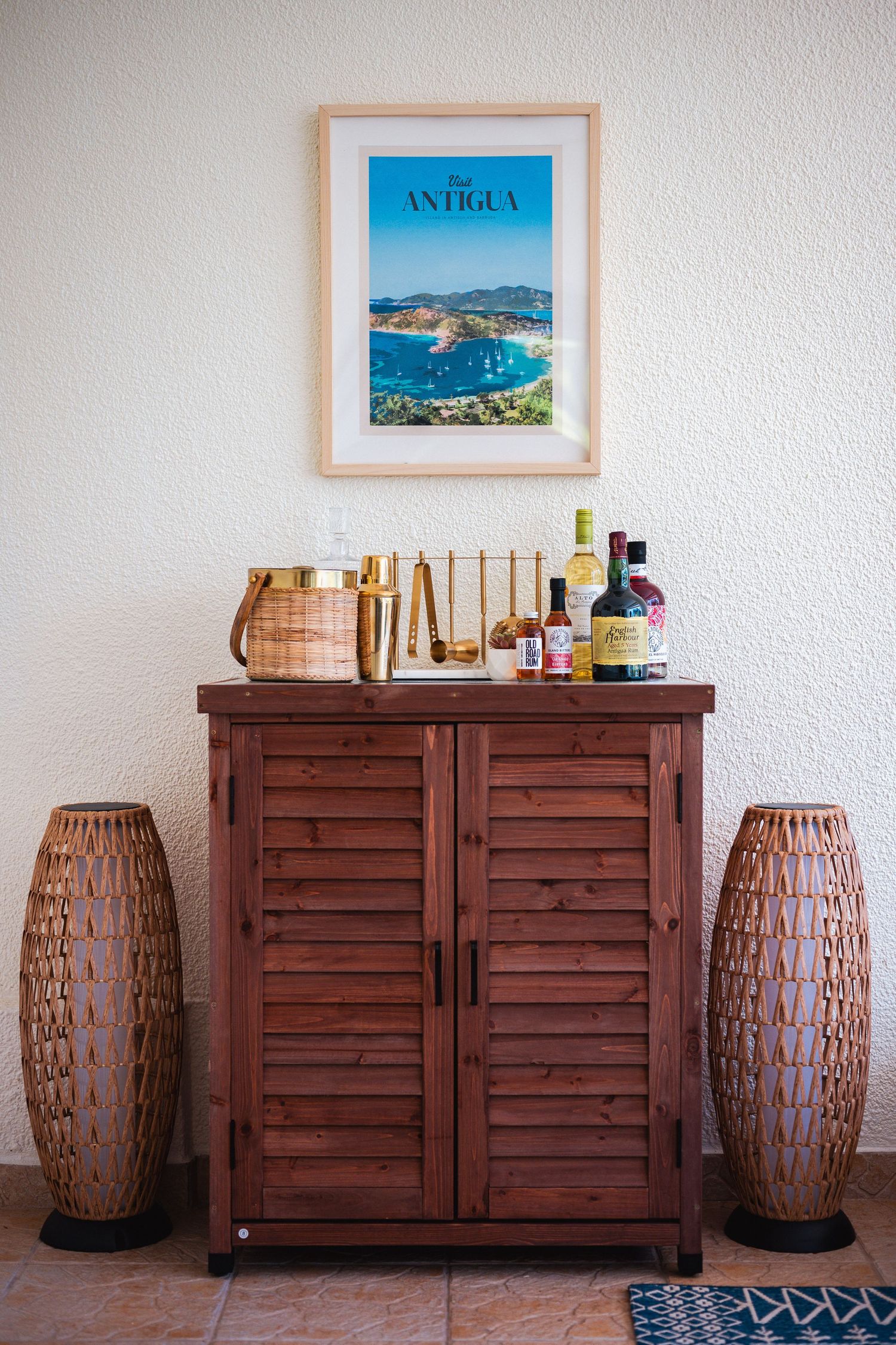 Wooden bar cabinet with liquor bottles beneath framed Antigua travel poster, flanked by woven rattan floor vases.