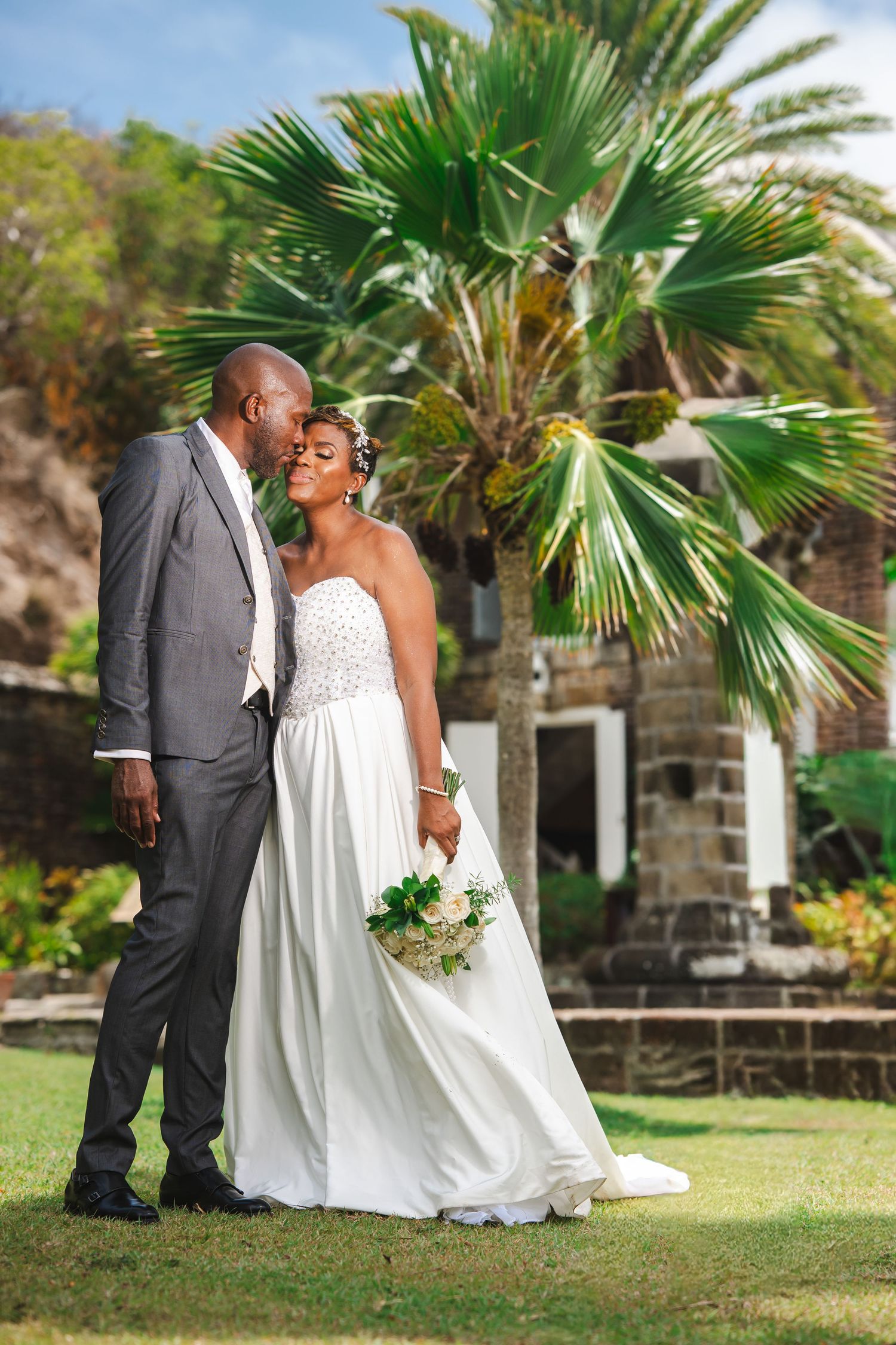A bride and groom share a tender moment outdoors, with palm trees and a stone building in a tropical setting.