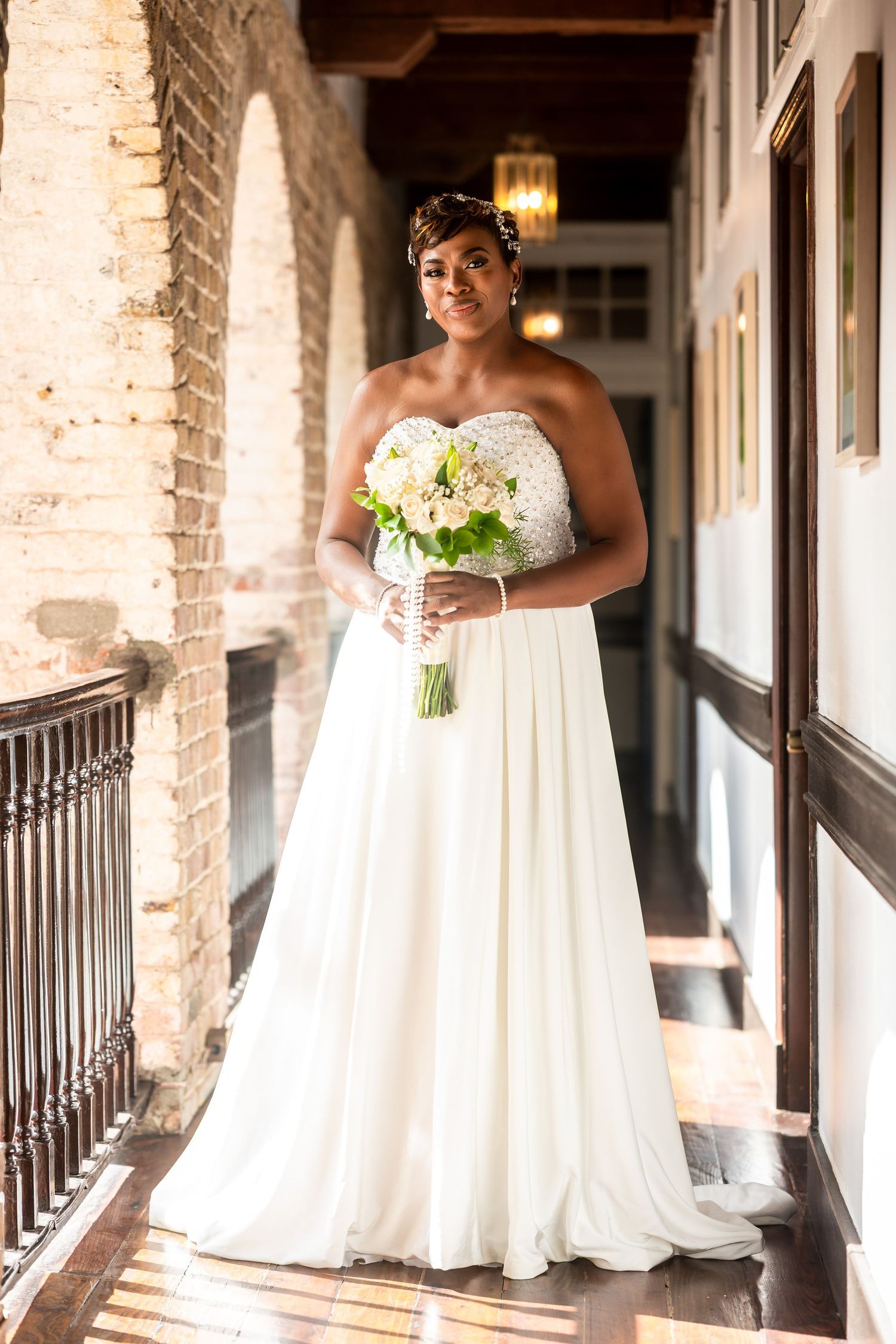 Bride in strapless white gown holding bouquet of white flowers on a sunlit historic venue balcony.