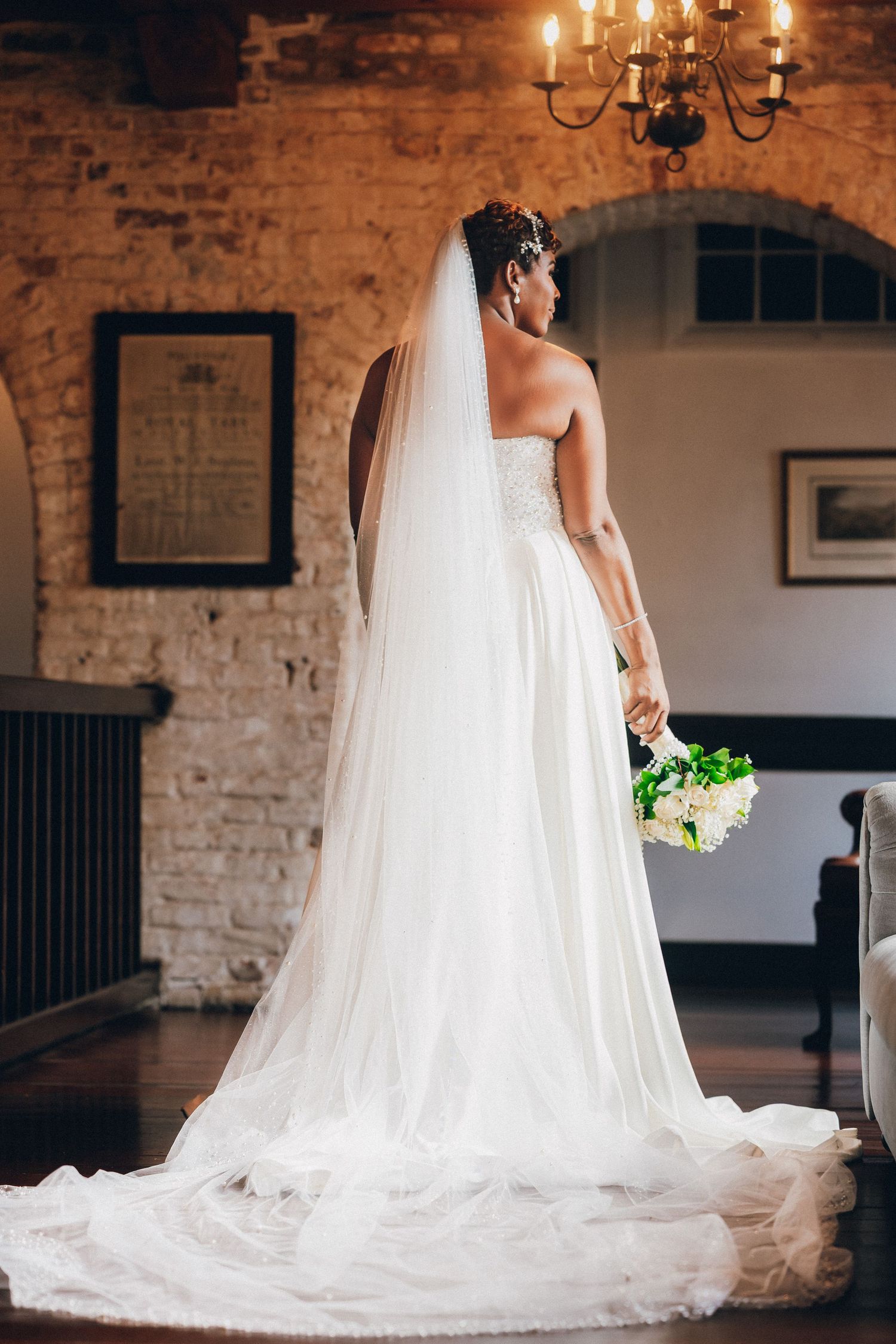 Bride in white strapless gown with long veil holds bouquet, standing in rustic brick venue with chandelier.