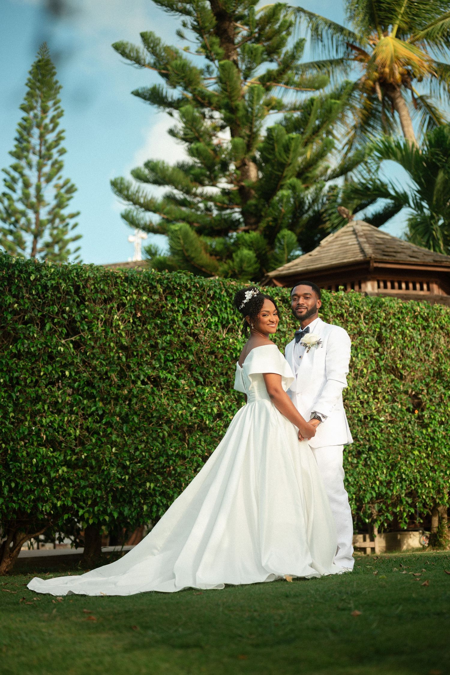 A bride and groom in white wedding attire pose hand-in-hand on a lush green lawn surrounded by tropical trees.