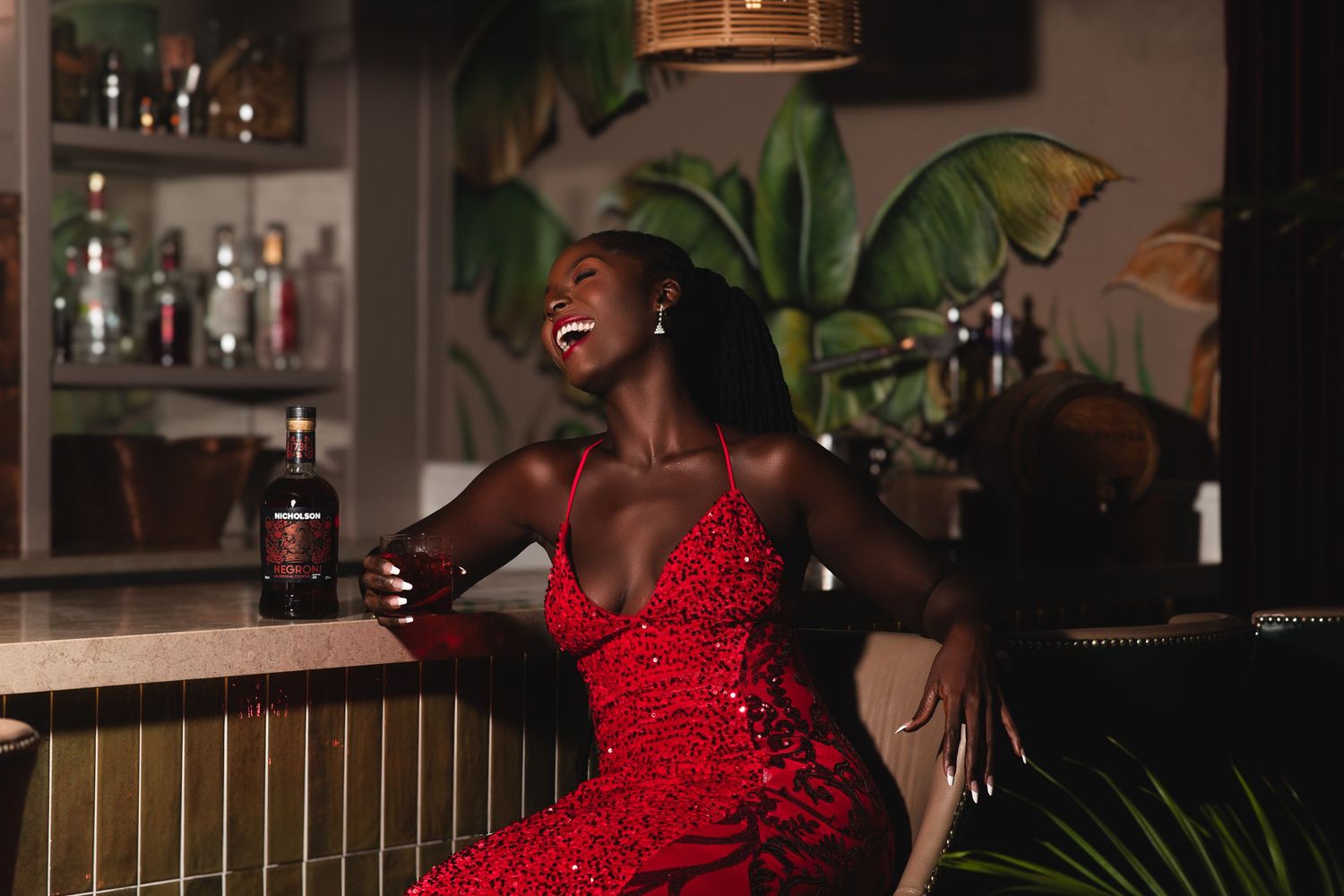 A joyful woman in a red sequin dress laughs at a bar beside a dark liquor bottle, surrounded by lush tropical decor.