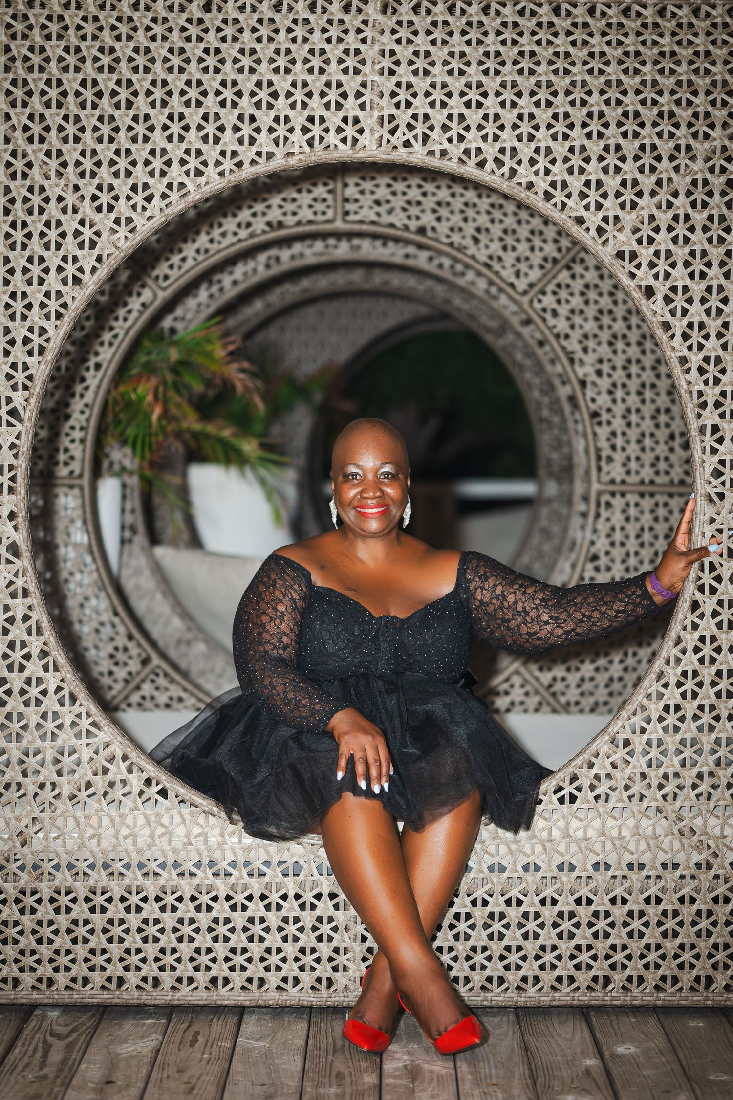 A smiling woman in a black lace dress and red heels sits in a circular decorative wall opening.