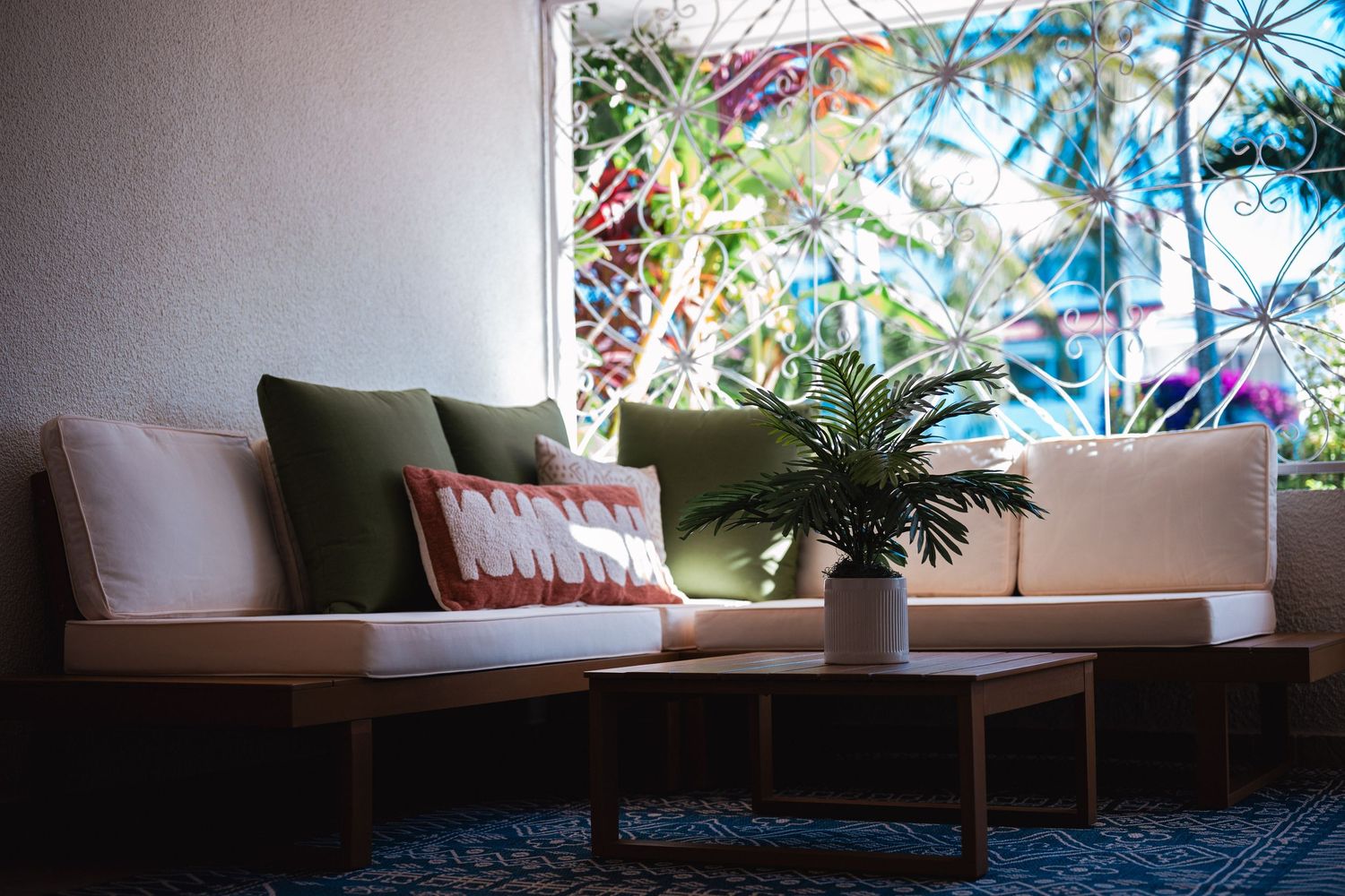 Tropical outdoor seating area featuring a beige sectional sofa with green pillows and potted palm on wooden coffee table.