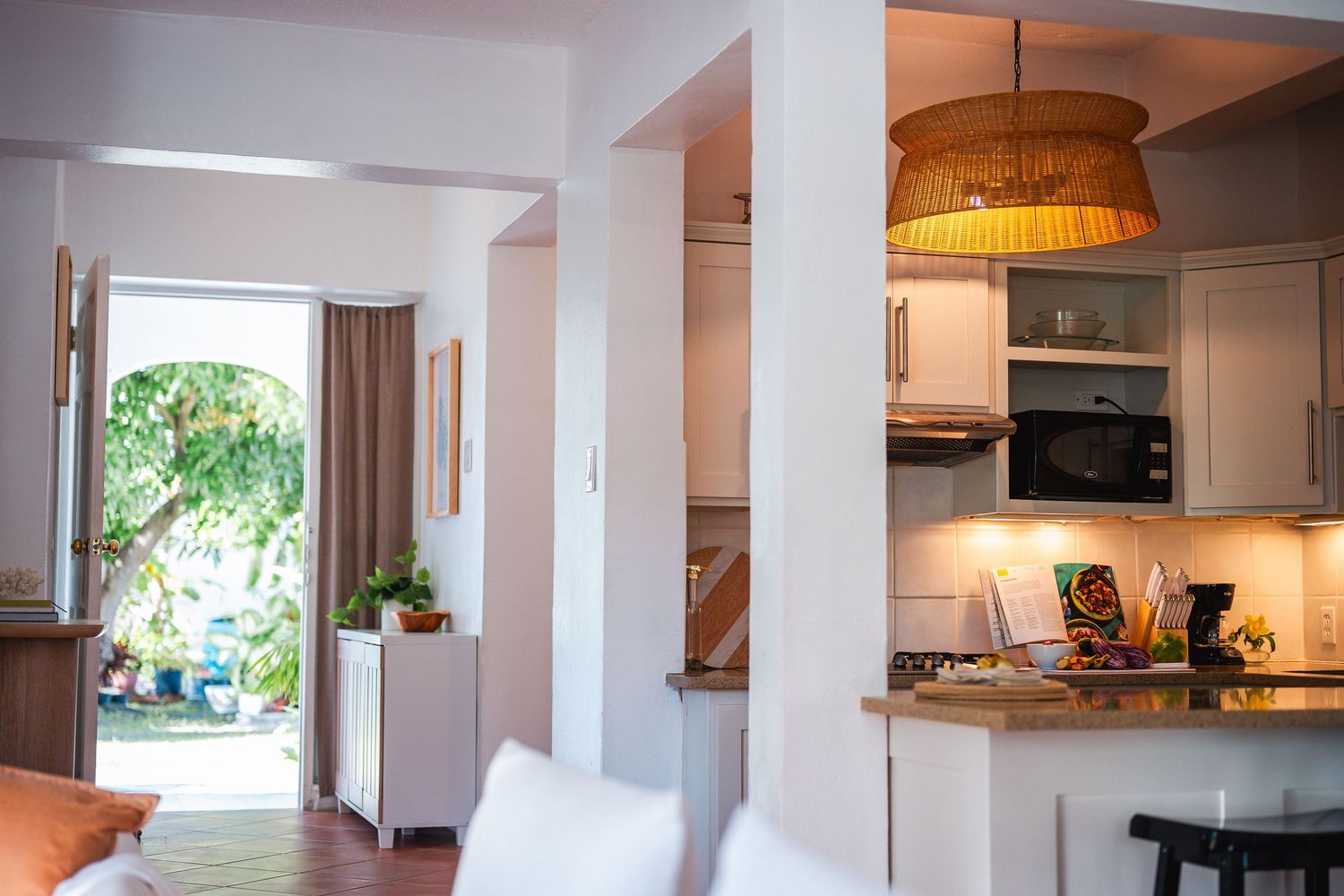 Bright open-plan kitchen and living area with white cabinets, wooden pendant light, and tropical garden view through doorway.