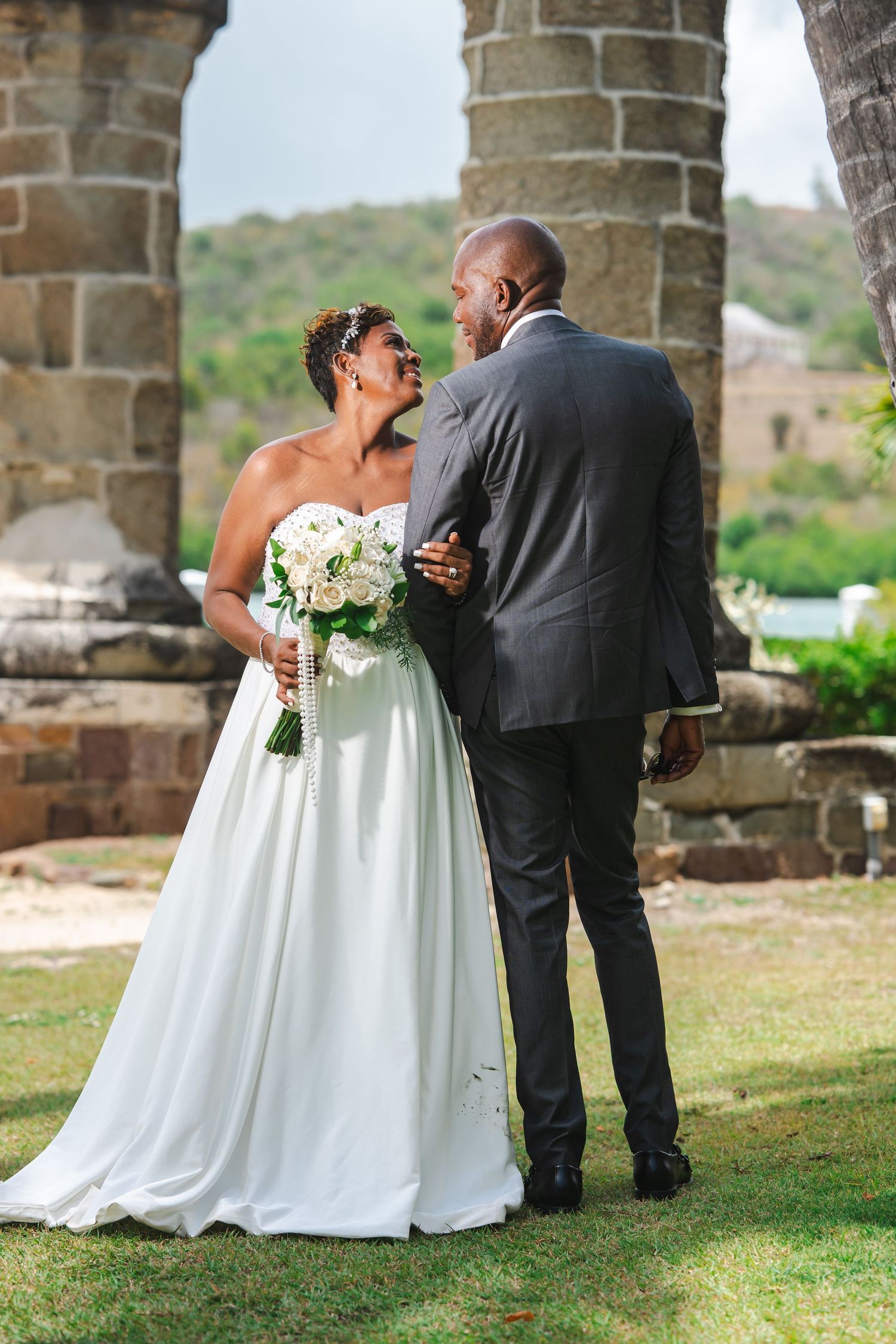 Bride and groom sharing a romantic moment outdoors near stone arches, bride holding white bouquet in wedding dress.