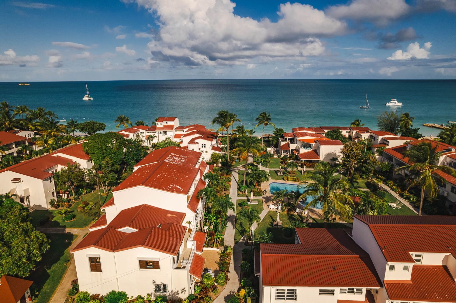 Aerial view of a Caribbean resort with red-roof villas, lush gardens, pool, and turquoise ocean backdrop.