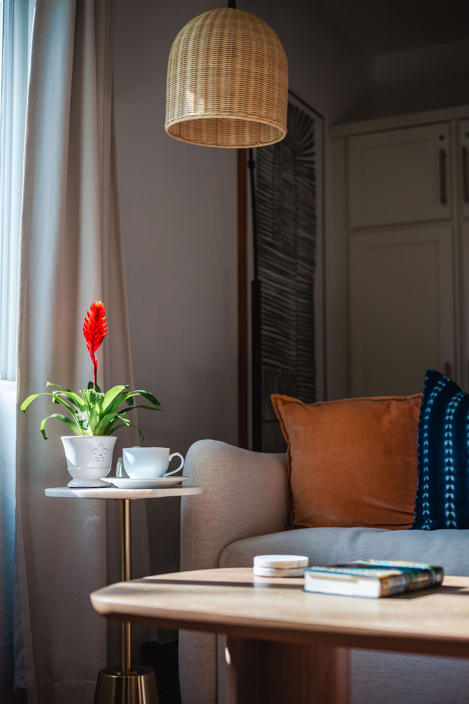 Cozy living room corner with wicker pendant lamp, red flower plant, and orange throw pillow on gray sofa.