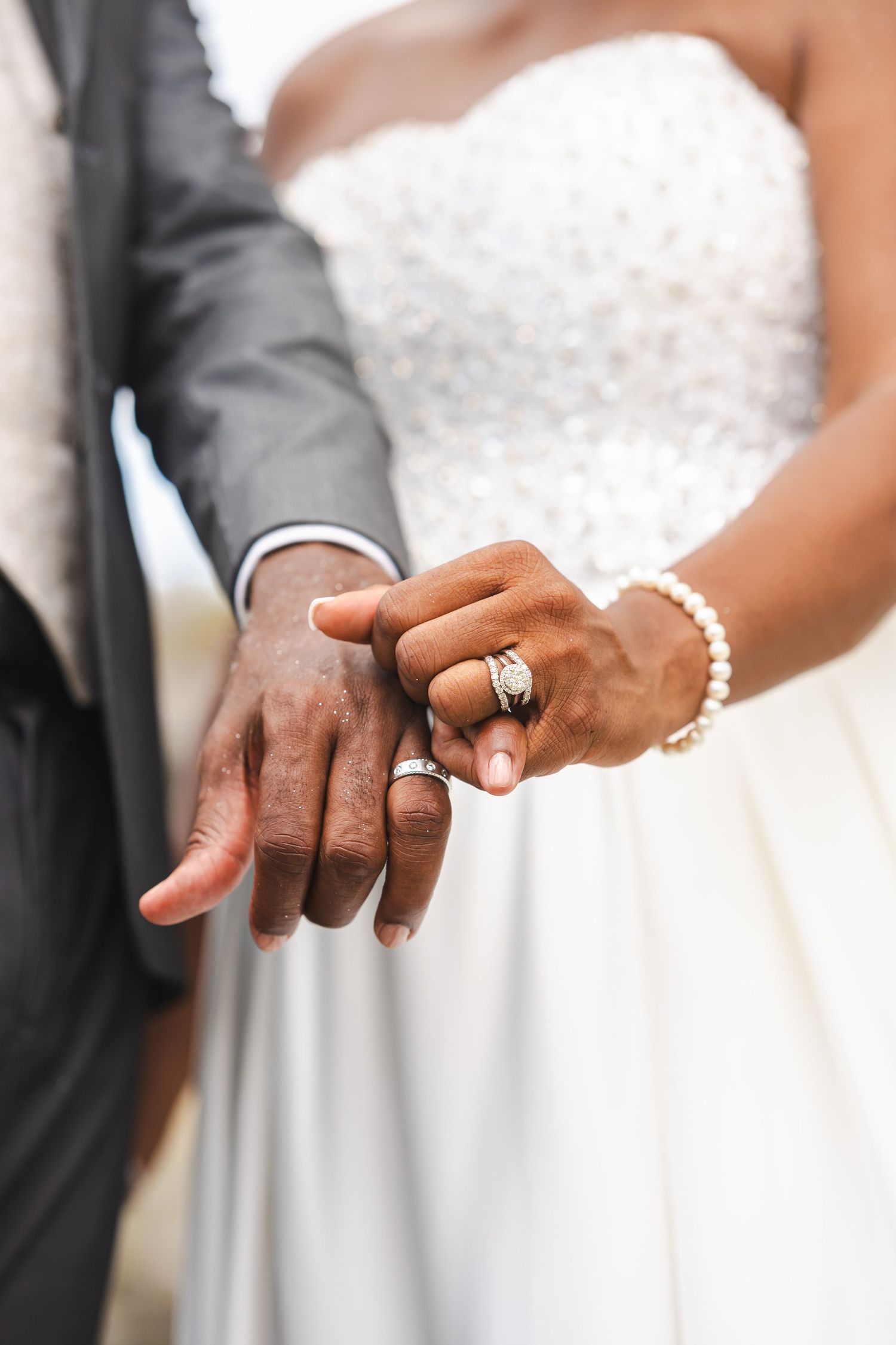 Close-up of newlywed couple holding hands, showcasing wedding rings, bride in white gown with pearl bracelet.