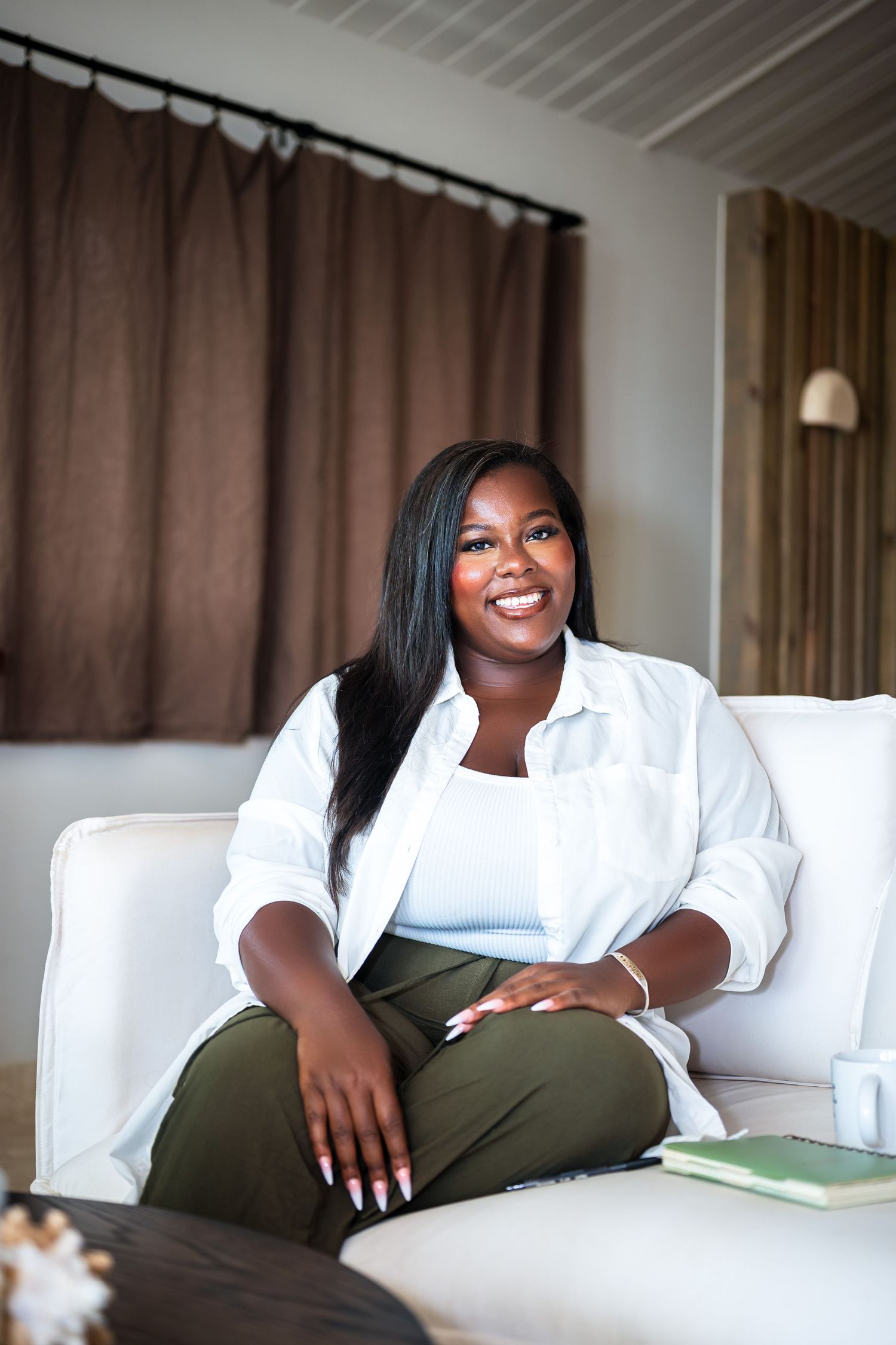 Smiling Black woman in white shirt and olive pants sitting cross-legged on a white sofa in a modern living room.