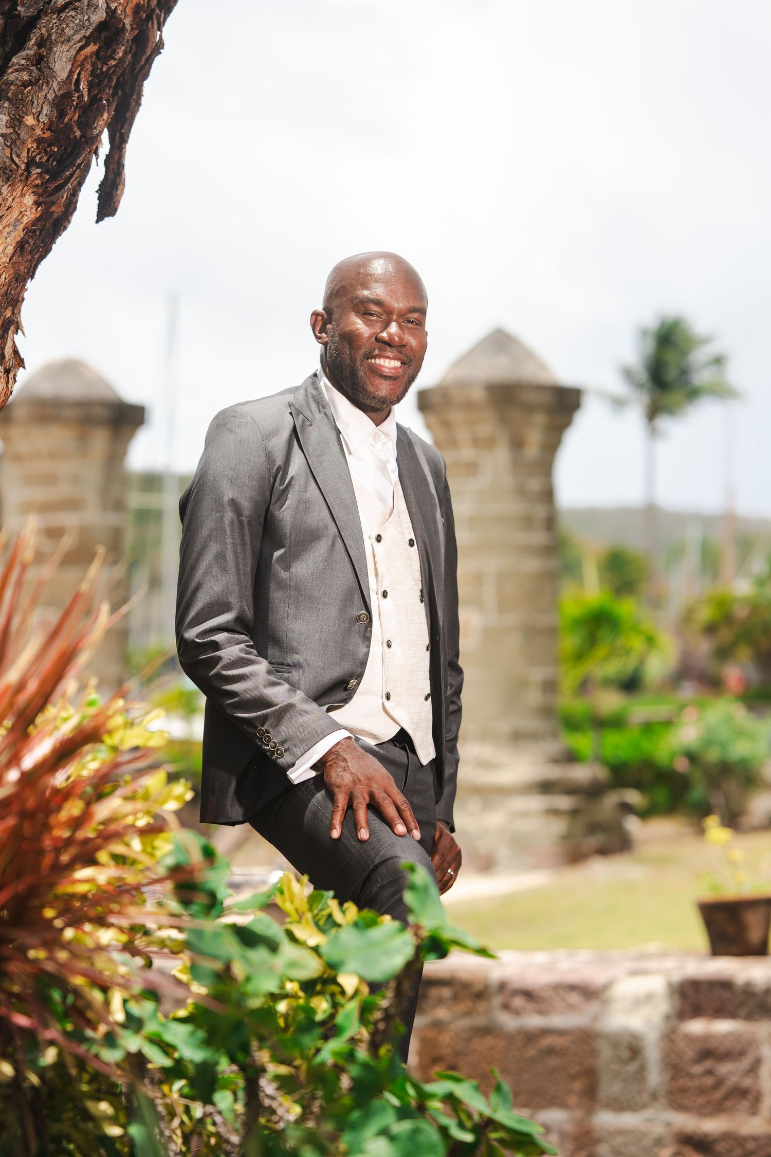 Smiling man in gray suit and white vest posing outdoors near tropical stone structures and lush greenery.